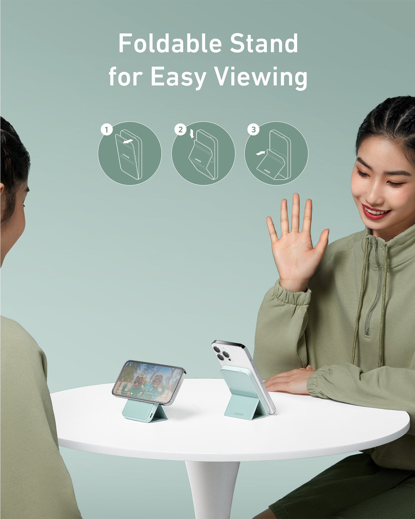 Two women sit at a round white table, each with a smartphone propped on an Anker 622 Magnetic Battery (MagGo) stand. One waves while icons show stand usage. Text reads: Foldable Stand for Easy Viewing with Anker, against a pale green background.