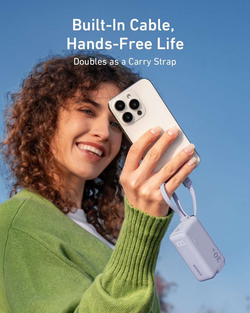 A smiling woman in a green sweater holds an Anker Power Bank (30W, 5k, Fusion, Built-In USB-C Cable) attached to her phone. Text above says: "Built-In Cable, Hands-Free Life. Doubles as a Carry Strap." Blue sky background.
