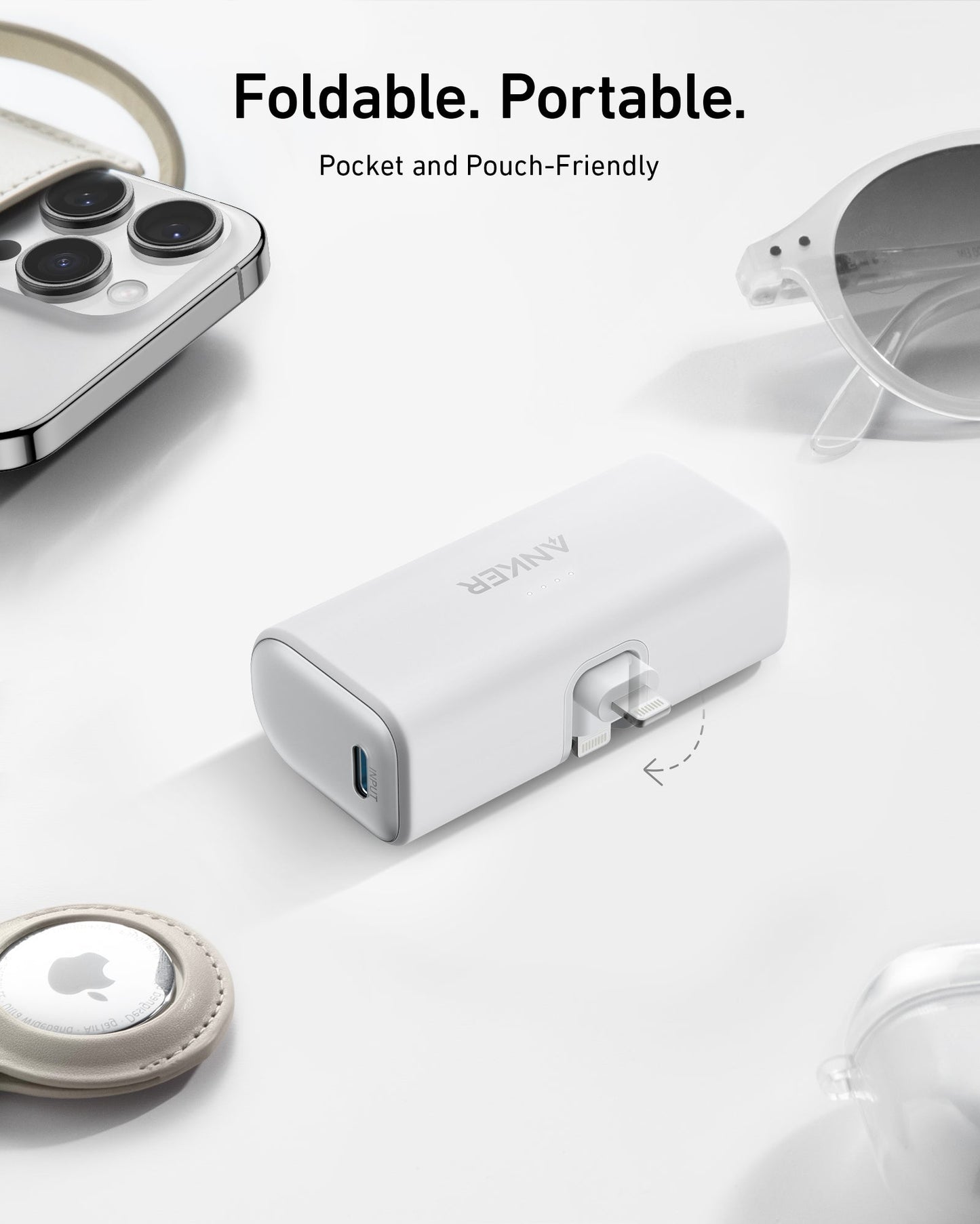 A compact, white Anker Nano Power Bank (12W, Built-In Lightning Connector) rests on a white surface alongside a smartphone, sunglasses, and a keychain with an Apple AirTag. Perfect for pairing with your MFi certified devices.