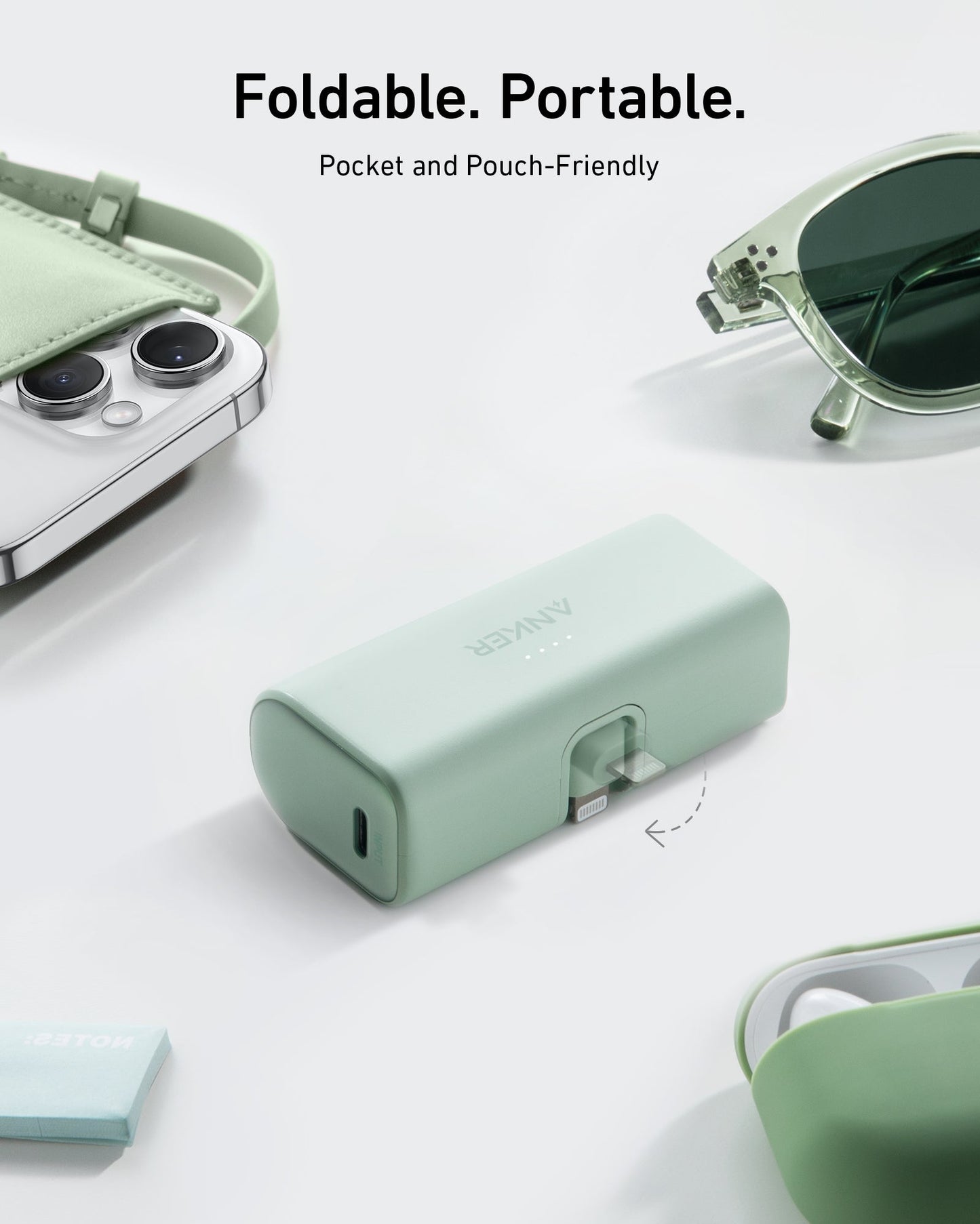 An Anker Nano Power Bank (12W, Built-In Lightning Connector) in mint green with a foldable plug rests on a light surface, surrounded by a matching phone case, sunglasses, earbuds case, and pouch. Text above says: "Foldable. Portable. Pocket and Pouch-Friendly.