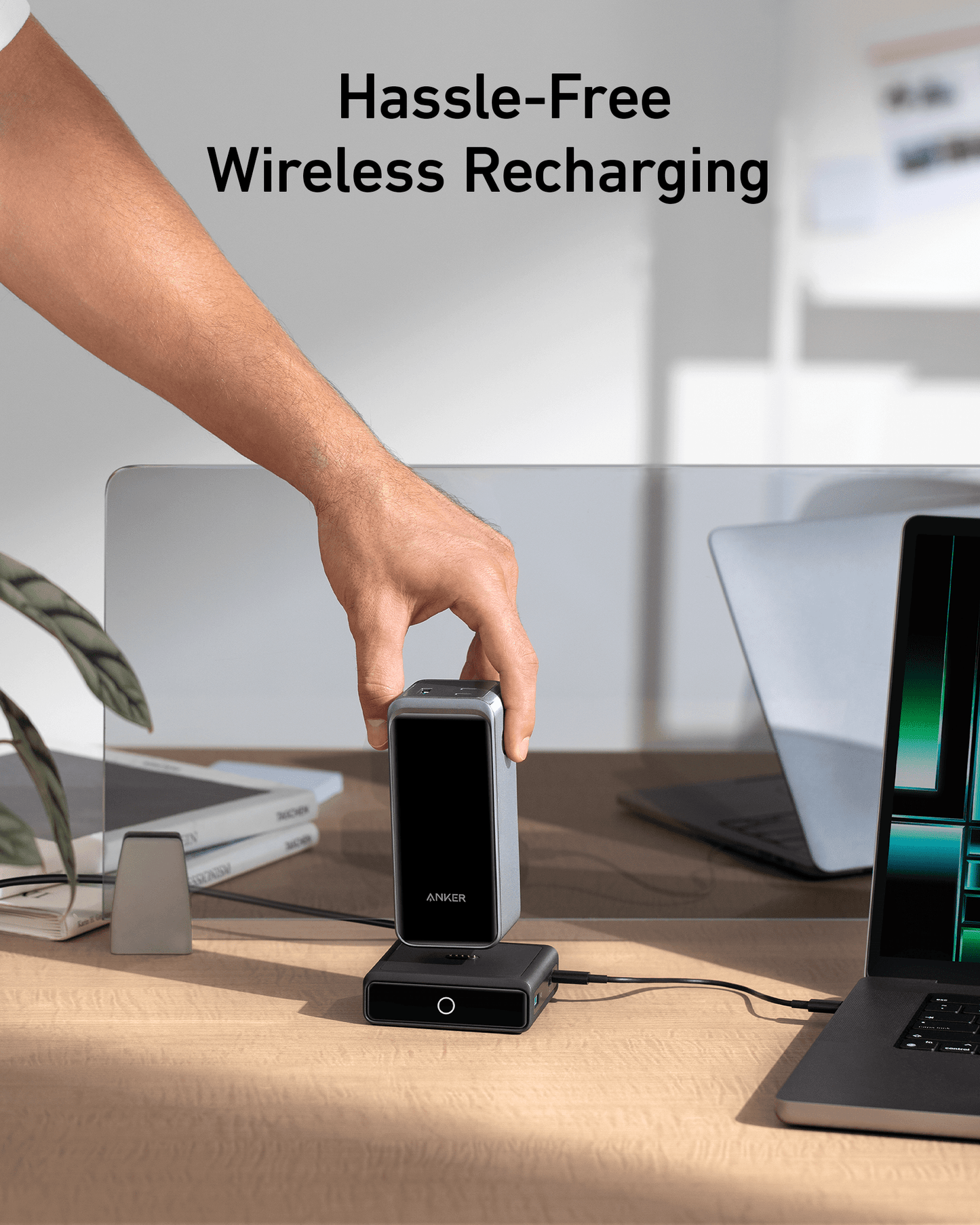 A hand places an Anker Prime Power Bank onto the Anker 100W Charging Base by Anker on a desk with a laptop, notebook, glasses, and potted plant nearby. “Hassle-Free Wireless Recharging” appears above, showcasing advanced GaN technology in a modern setting.
