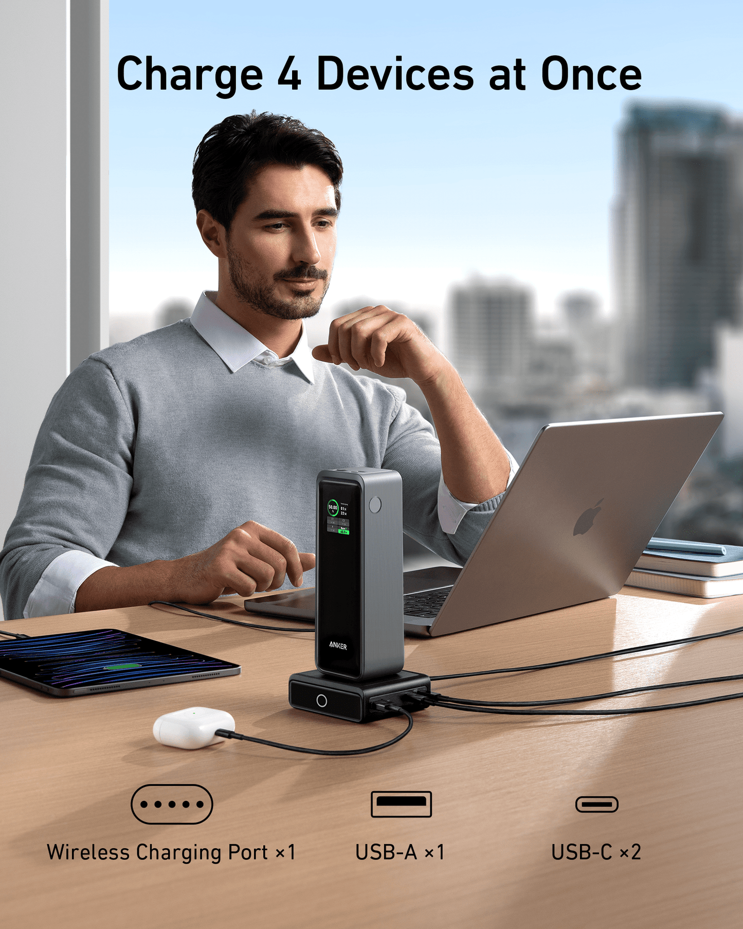 A man sits at a desk using a laptop, with the Anker 100W Charging Base for Anker Prime Power Bank powering his smartphone, tablet, and earbuds. Above him, text reads “Charge 4 Devices at Once,” with icons for wireless charging and GaN tech.