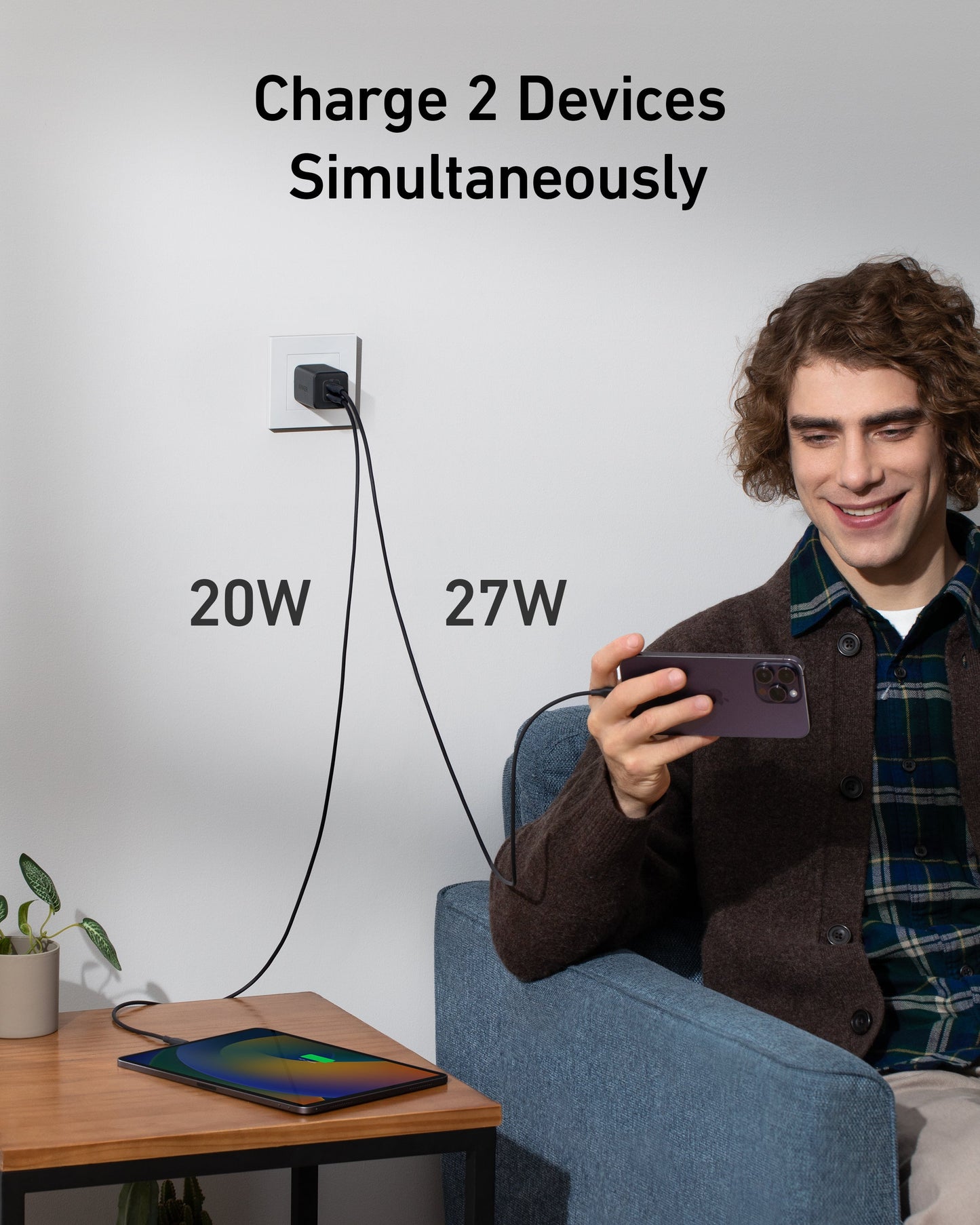A young man smiles on a sofa, holding his smartphone plugged into an Anker 523 Charger (Nano 3, 47W), while another cable powers a tablet. Above, text reads "Charge 2 Devices Simultaneously" with “20W” and “27W” labels.
