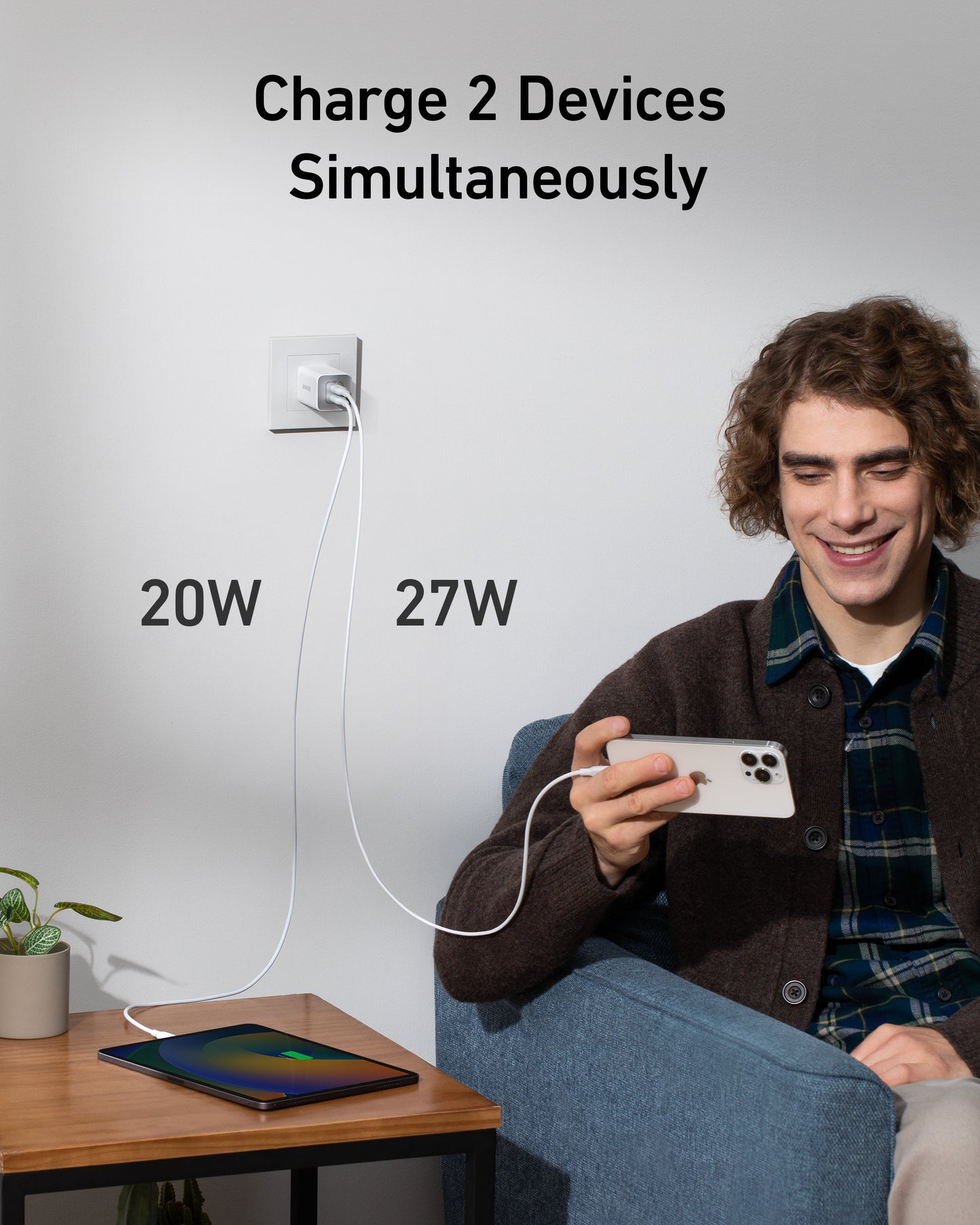 A young man smiles on a blue armchair, charging his phone with the Anker 523 Charger (Nano 3, 47W) while a tablet fast charges on a wooden table. Text: "Charge 2 Devices Simultaneously" with “20W” and “27W” by each device.