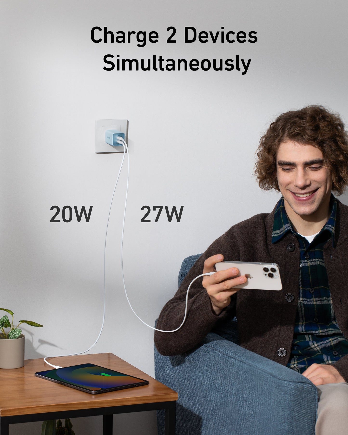 A young man, smiling at his phone as it fast charges with the Anker 523 Charger (Nano 3, 47W), relaxes in an armchair. Next to him, a tablet charges too. Text above: “Charge 2 Devices Simultaneously.”.