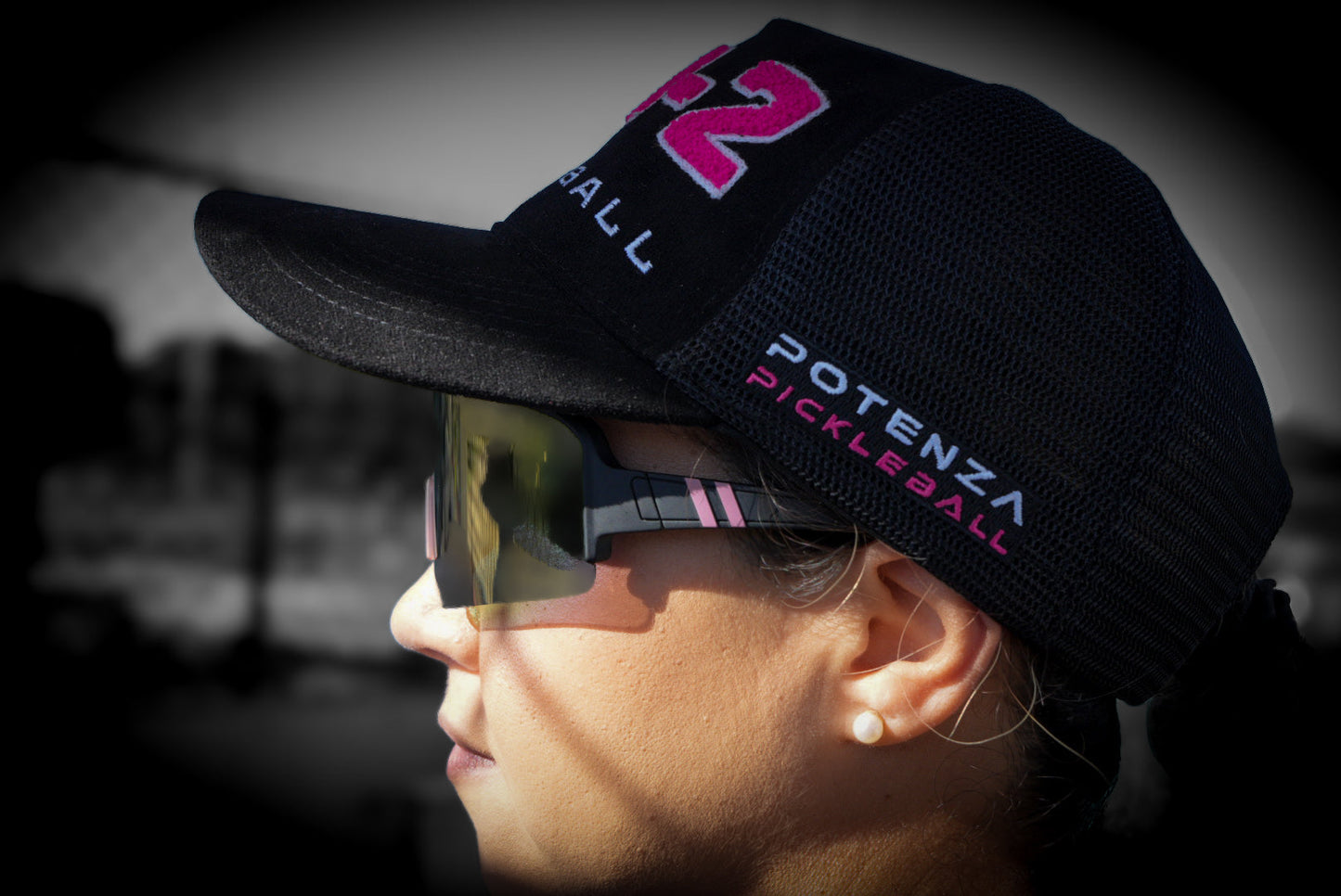 Close-up side view of a person wearing Potenza Pickleball’s "0-0-2" Collection Hat (0-0-START) with embroidered chenille letters, plus dark reflective sunglasses. A blurred background and soft vignette highlight the hat.
