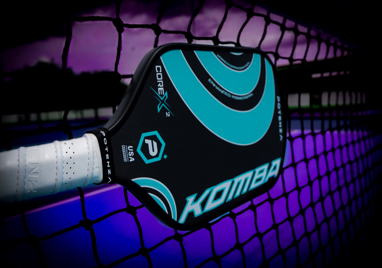 A KOMBA+ COREx2 carbon fiber pickleball paddle by Potenza Pickleball, featuring a black design with teal accents and a white grip, leans against a net on a purple and blue court at dusk under a dramatic sky.