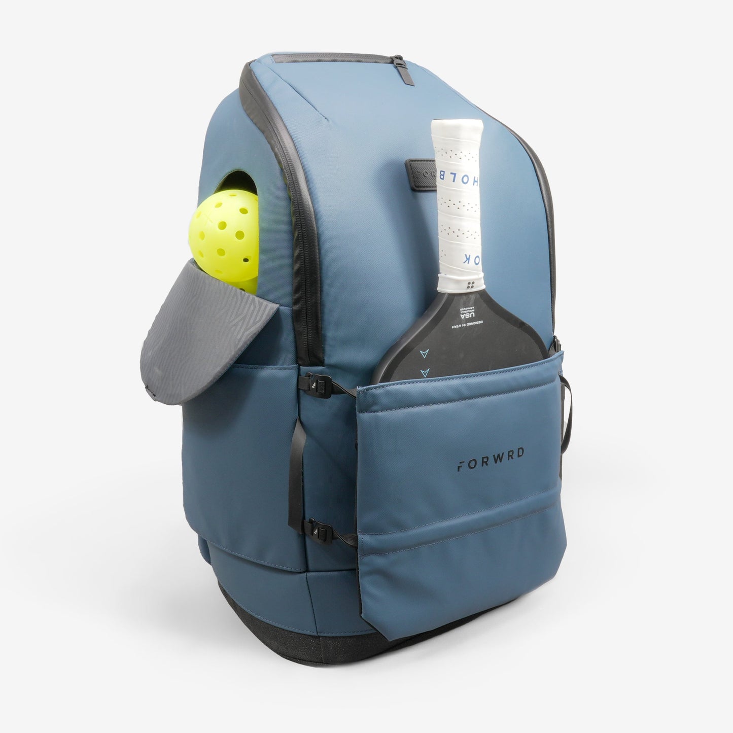 A blue backpack with a black base stands upright. The FORWRD Court Caddy Front Paddle Sleeve holds a pickleball paddle in its labeled front pocket, while two yellow balls peek from a side pocket beneath a gray flap.