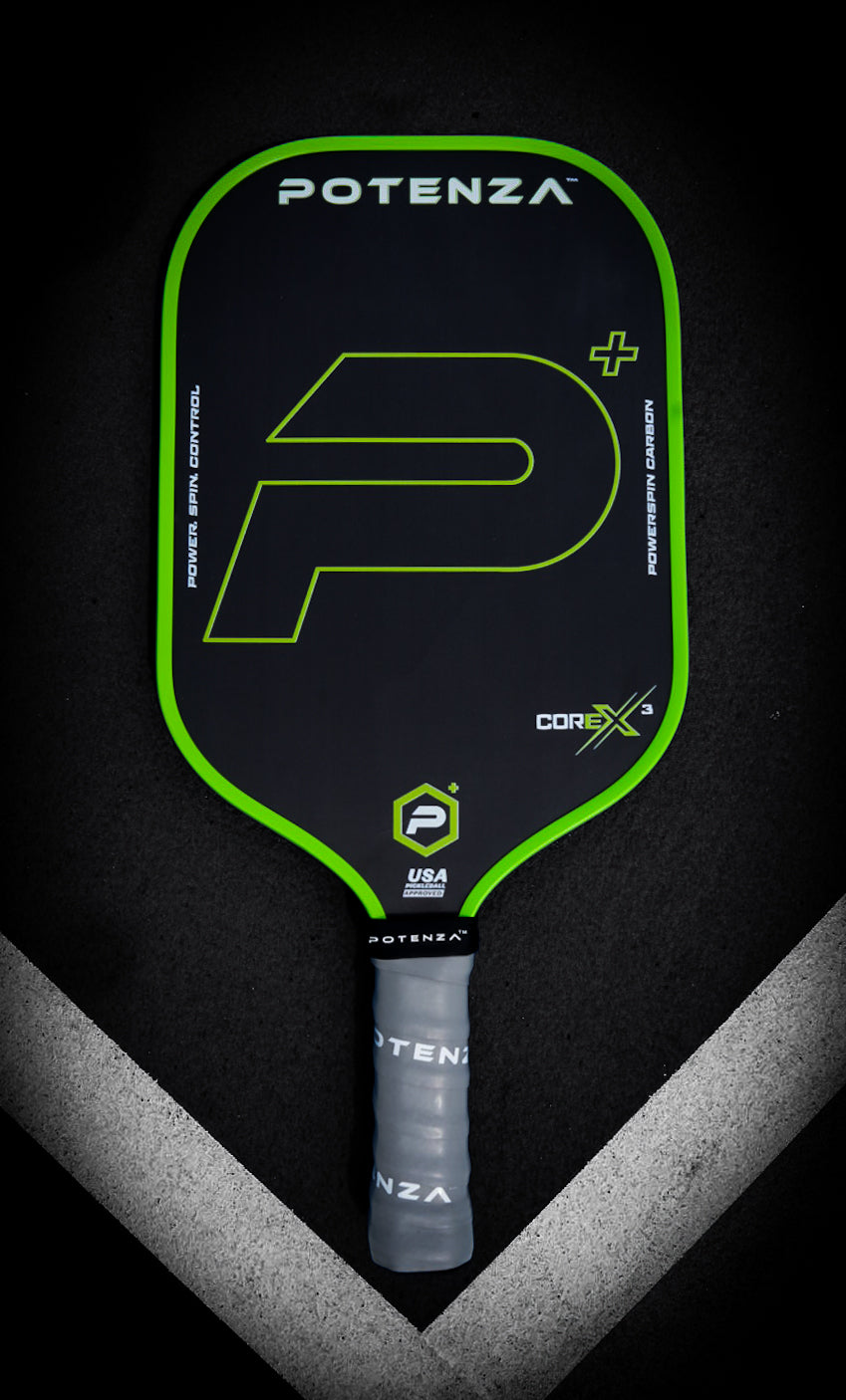 A black Potenza Pickleball P+ PowerSpin Carbon COREx3 paddle (Neon Green, Elongated Face) with neon green edges, a large green “P”, and a gray grip rests on a dark surface crossed by two white court lines.