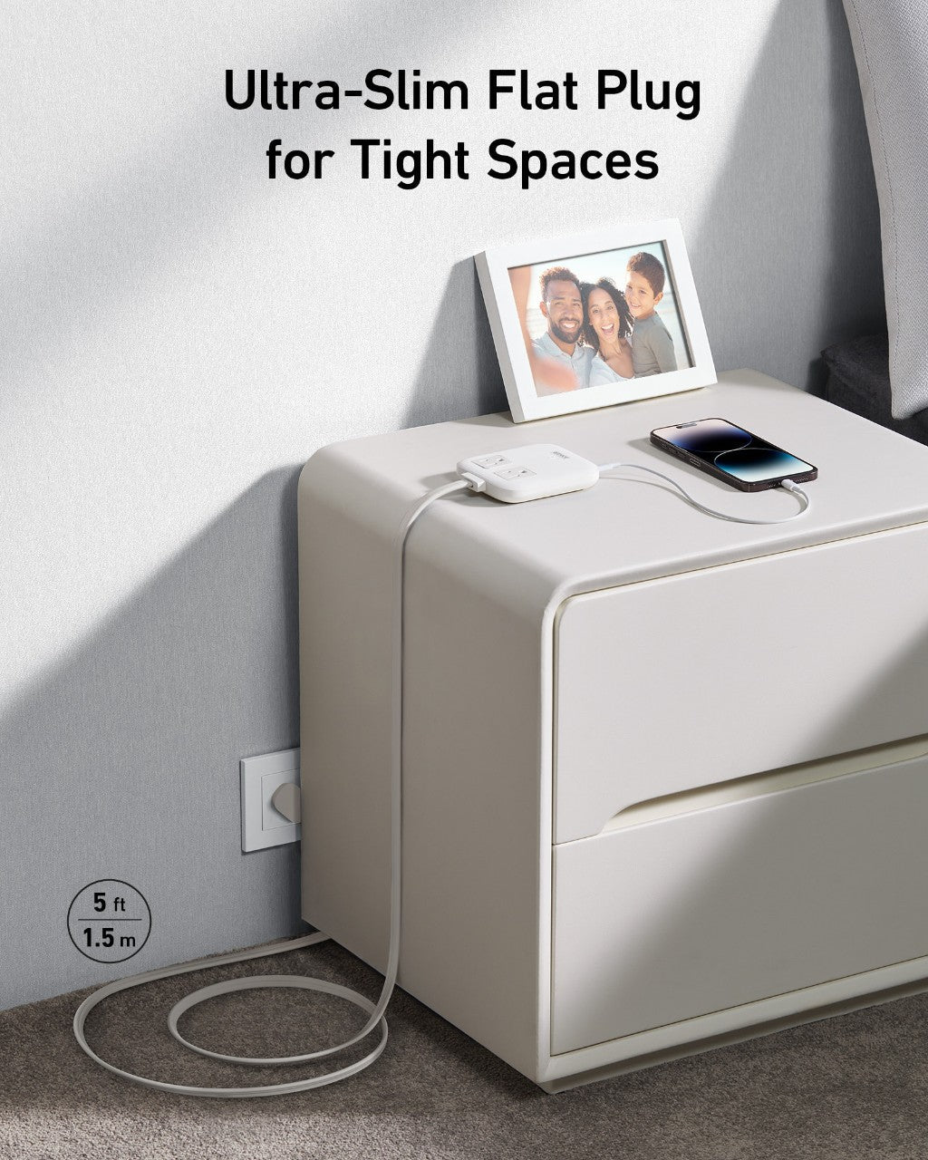 An Anker Nano Charging Station (6-in-1, 67W) sits on a white nightstand, charging a smartphone and powering devices with its USB-C ports. Its ultra-slim 5 ft (1.5 m) flat plug extension cord reaches the outlet. Nearby, a framed family photo. Ultra-Slim Flat Plug for Tight Spaces.