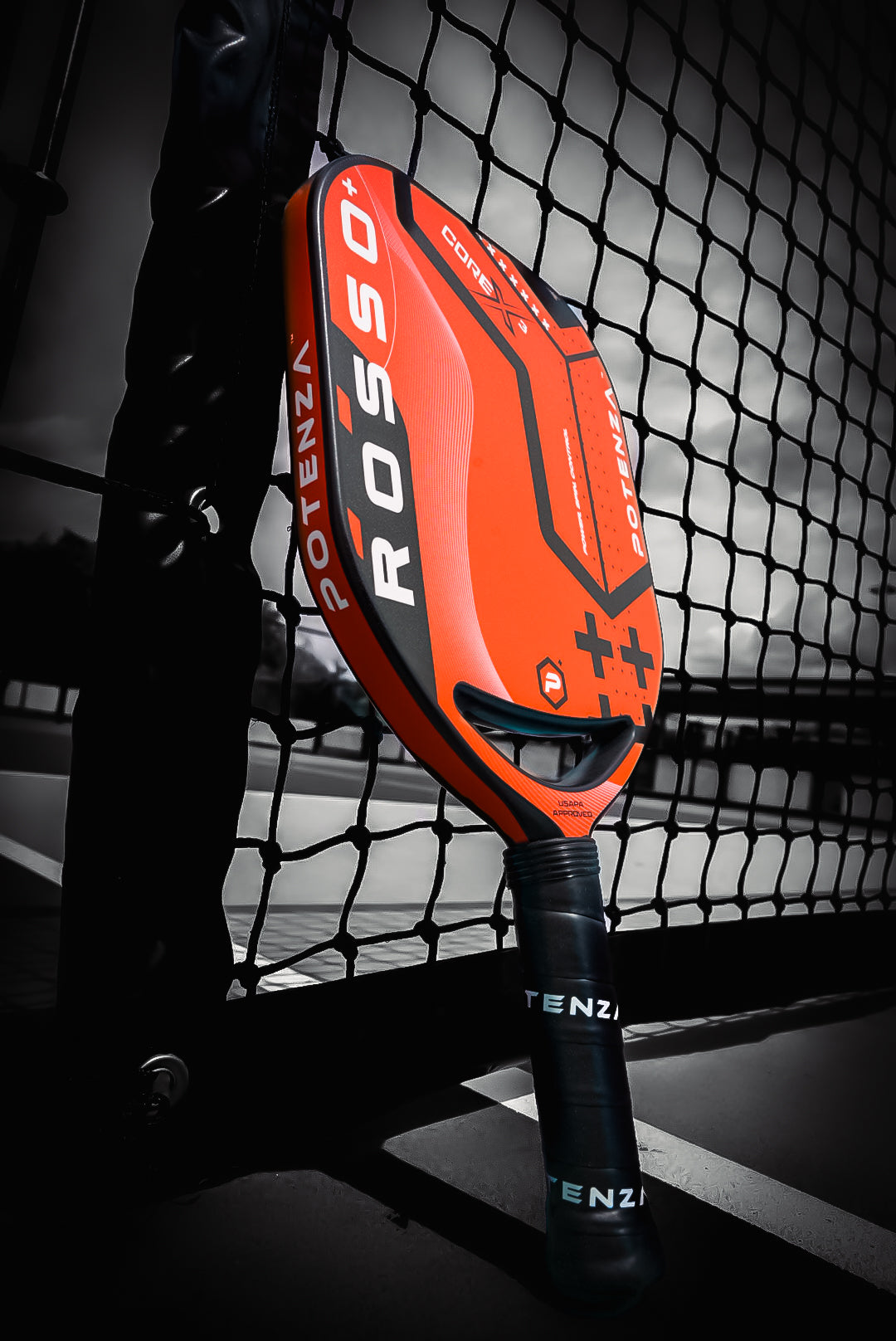 The Potenza Pickleball ROSSO+ COREx3 paddle, with a bold 21mm grip, leans against an outdoor net. Its red and black design stands out in vivid color against the mainly black and white court.