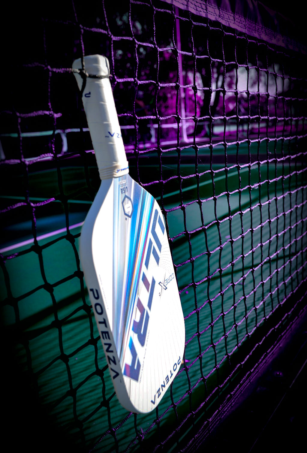 A white Potenza Pickleball ULTRA+ COREx2 paddle with colorful stripes leans against a black net on an outdoor court, bathed in deep shadows and purple light. "ULTRA" and "POTENZA" are printed on the paddle.