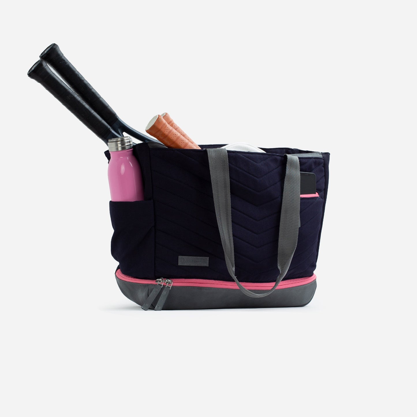 The Doubletake PARISII Pickleball Tote in navy and gray, featuring gray handles, pink accents, and a front pocket, holds two black bats, a pink water bottle, and a brown glove on a white surface with a light gray background.
