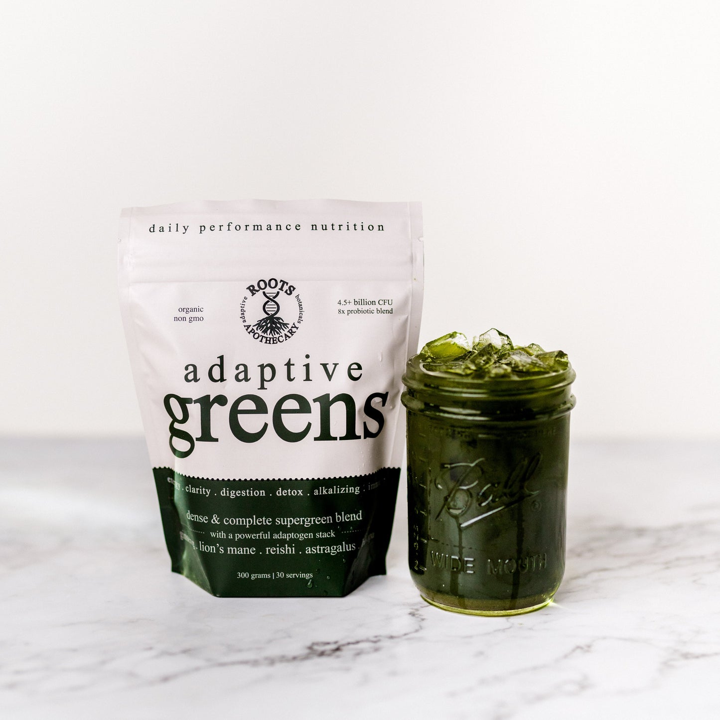 A white packet labeled "adaptive greens. performance superfood." by Roots Apothecary stands beside a mason jar with green liquid and ice on a white marble surface, highlighting “daily performance nutrition,” “organic,” and adaptogens like lion’s mane, reishi, and astragalus.