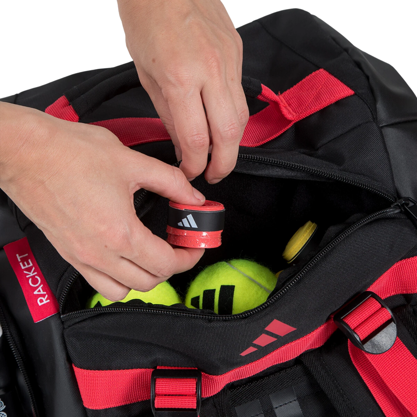 A close-up of hands placing a red tennis racket grip into the spacious Adidas TOUR BLACK Racket Bag 3.4 by Padel USA, alongside two yellow tennis balls and another grip. A red zipper pull labeled RACKET is visible on the bag's left side.