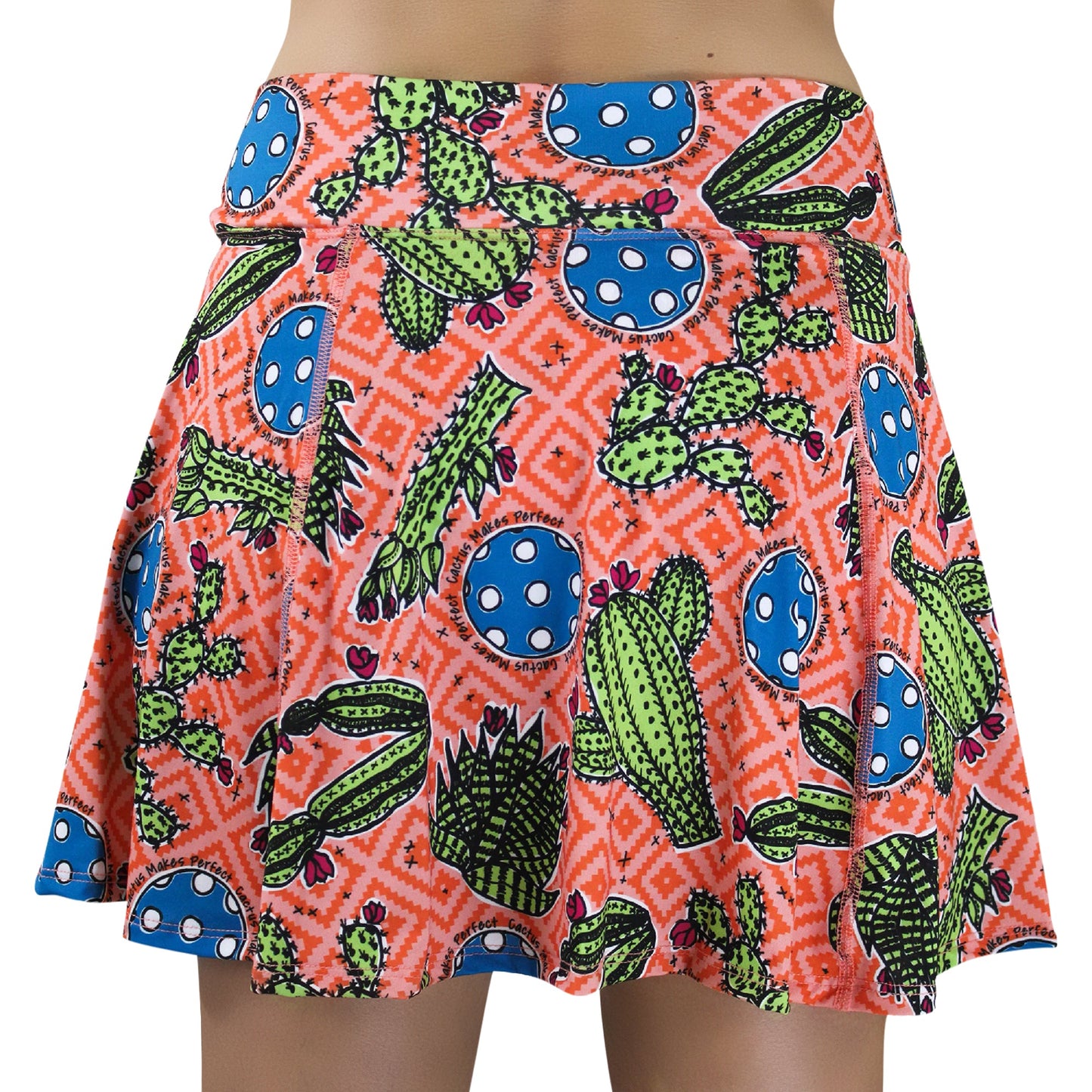The Pickleball Bella PB Aline Skort Cactus Makes Perfect 1 features a bold cactus, blue circle, and white spot print on orange with geometric details, built-in compression shorts, and a waist-to-mid-thigh fit.