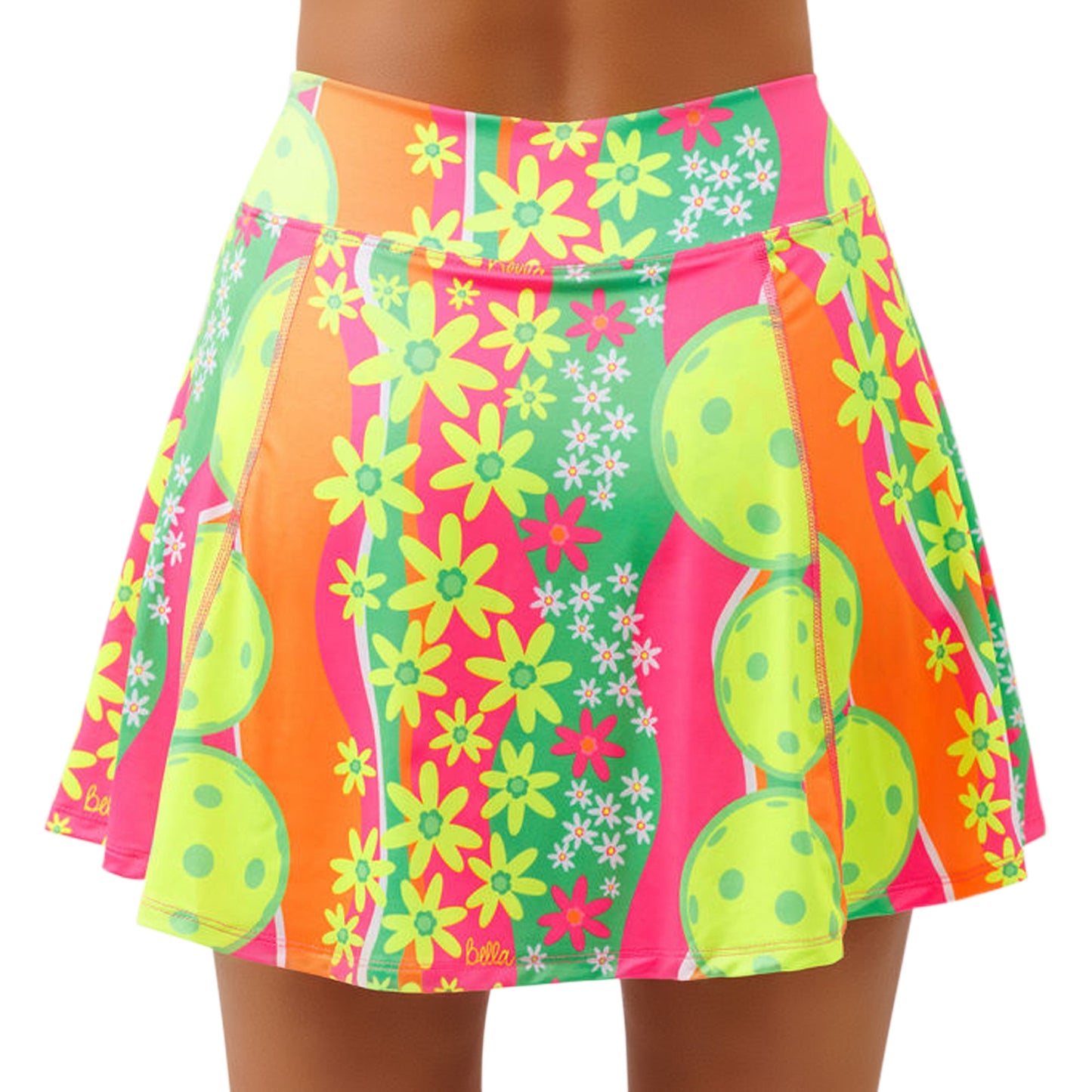 The PB Aline Skort Daisy by Pickleball Bella features a vibrant pattern of yellow flowers, lime-green pickleball paddles and balls, and wavy orange, pink, and green stripes on 4-way stretch fabric. Shown flaring on a medium-brown-skinned model.