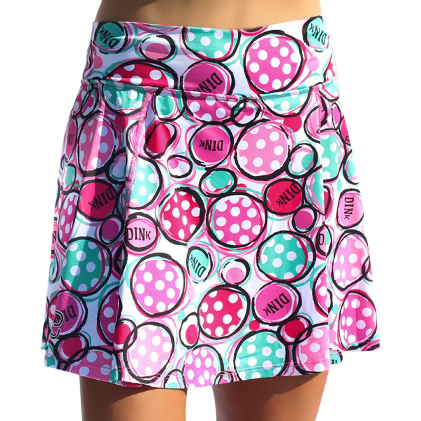 A woman wears the Pickleball Bella PB Aline Skort Dink 2, a colorful pickleball skirt with overlapping pink, turquoise, and white circles—some with polka dots, others with “DINK.” It features a wide waistband and built-in compression shorts.