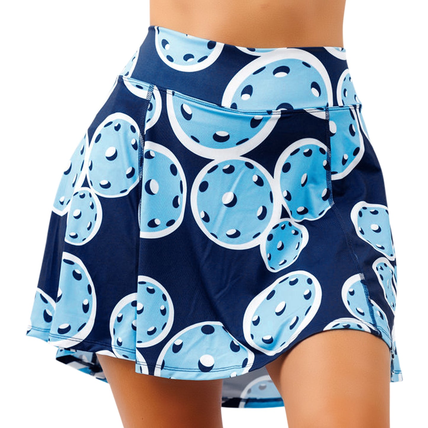 A woman models the Pickleball Bella PB Aline Skort Double Bounce in navy, featuring playful blue pickleballs with white details and a flared hem. The smooth 4-way stretch fabric offers comfort and flexibility. Only her midsection and upper legs are visible.