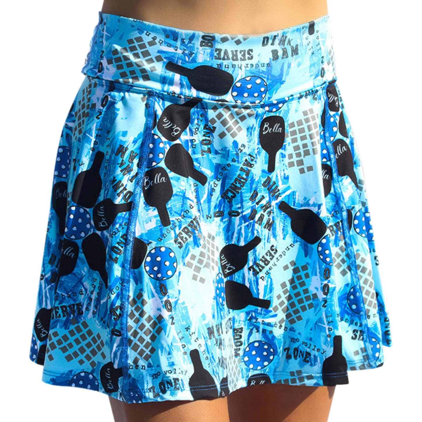 A person wears the Pickleball Bella PB Aline Skort Graffiti 2, a blue athletic skirt with black paddle silhouettes, blue polka-dot balls, and words like “Bella” and “Serve” over abstract grid and splash patterns.