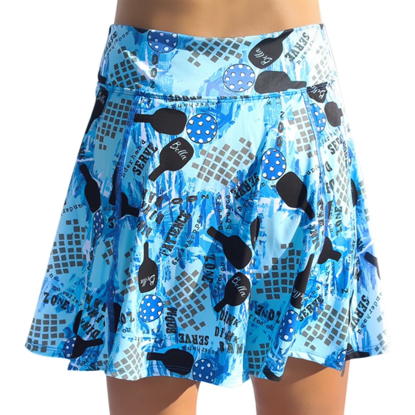 A person wears the Pickleball Bella PB Aline Skort Graffiti 2, featuring black and blue polka-dot paddles, white text, and abstract grids. The flared athletic skort stands out on a white background with its fun, sporty print.
