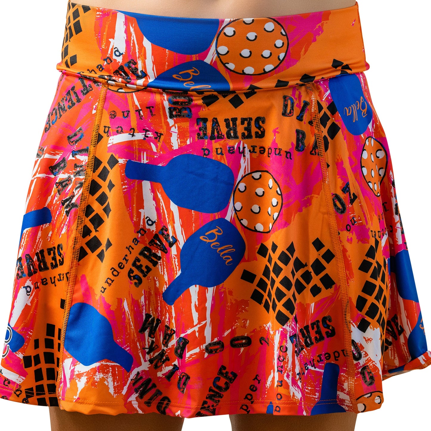 The Pickleball Bella PB Aline Skort Graffiti 3 features a bold abstract design with blue paddles, orange balls with white dots, and black text accents on an orange background with pink and white splatters in a stylish A-line cut.
