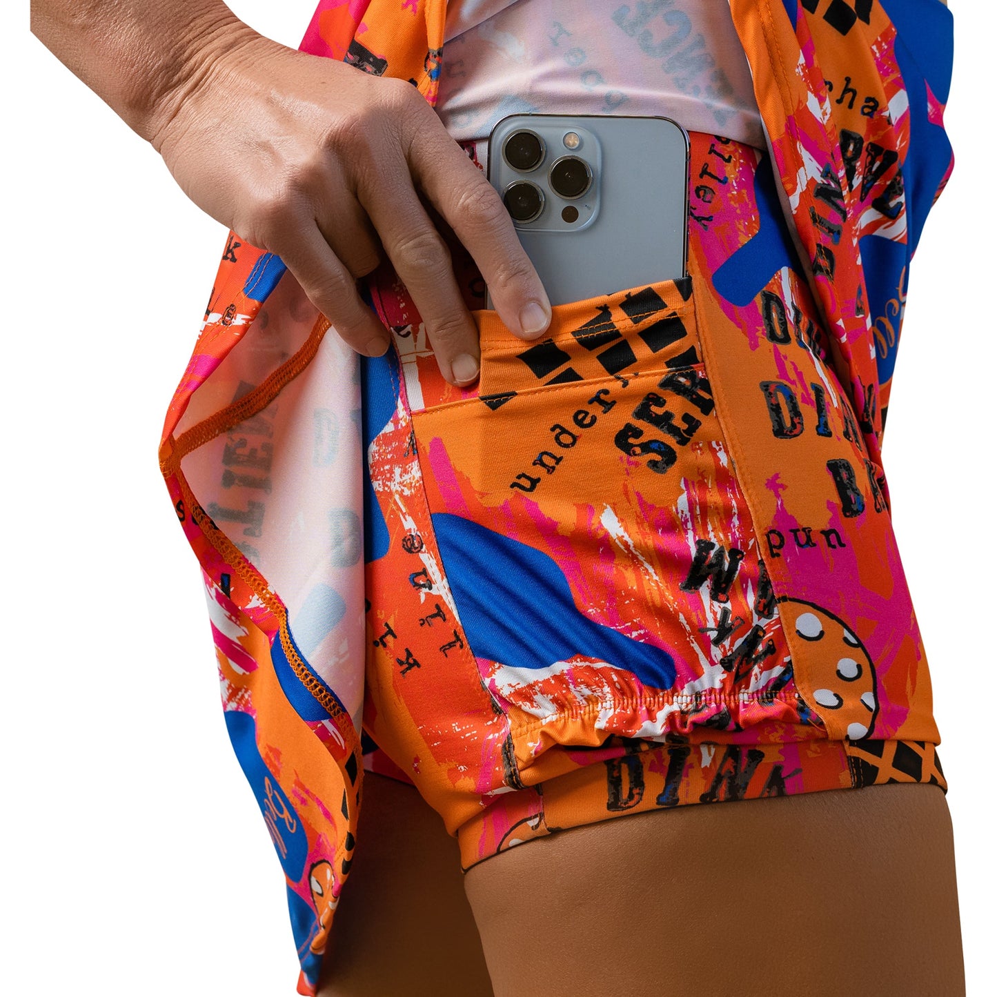 A close-up shows a light-brown hand sliding a silver smartphone into the side pocket of Pickleball Bella’s PB Aline Skort Graffiti 3, featuring bright orange and blue patterns with bold black text. The shirt matches the skort.