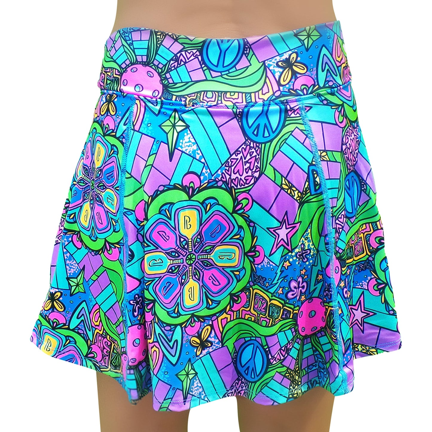 The PB Aline Skort Groovy by Pickleball Bella features a bold, psychedelic retro print in blue, green, pink, and purple with large flowers, peace signs, stars, and geometric shapes. This flared skort sits high on the waist for playful vintage style.