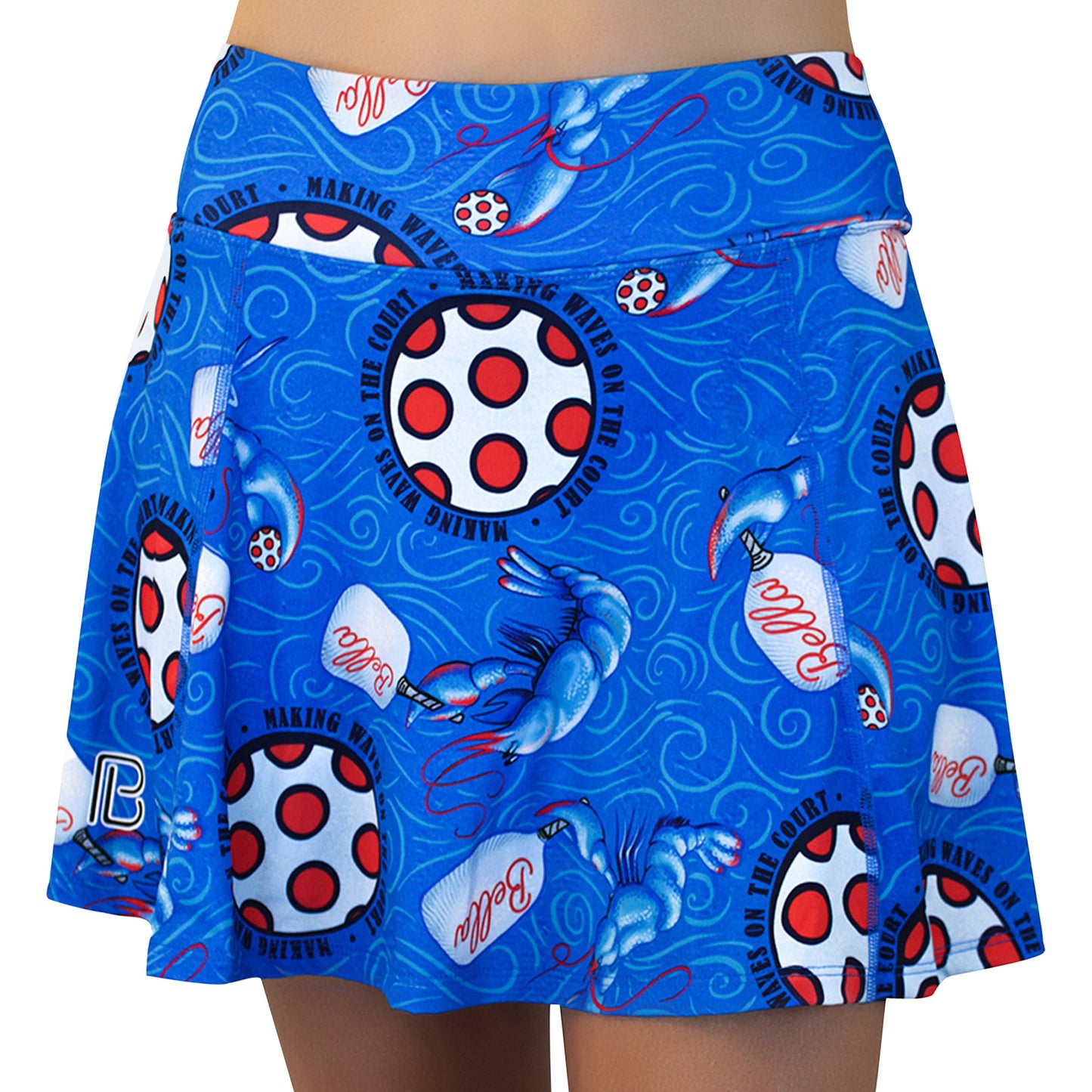 Pickleball Bella's PB Aline Skort Lobster Monster is a blue athletic skort with red and white pickleballs, blue paddles, "Dink" script, and "Making Waves on the Court" in black circles. Modeled by a person with light skin.