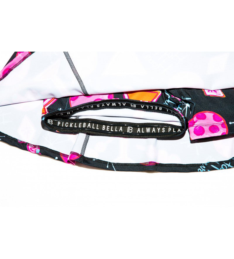 Close-up of the PB Aline Skort Martini 2-4-1 Happy Hour by Pickleball Bella, featuring a waistband with vibrant pink, orange, and purple shapes on black. The inner band has white PICKLEBALL BELLA text on black and a white fabric lining.