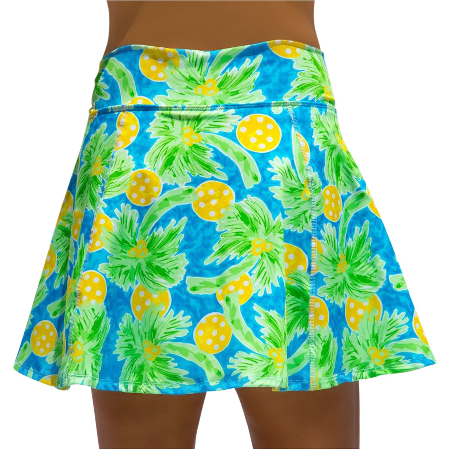A person wearing the Pickleball Bella PB Aline Skort Palms 1—a blue skater skirt with green palm leaves, yellow polka-dotted fruits, a wide waistband, discreet compression shorts, and a flared fit above the knee.