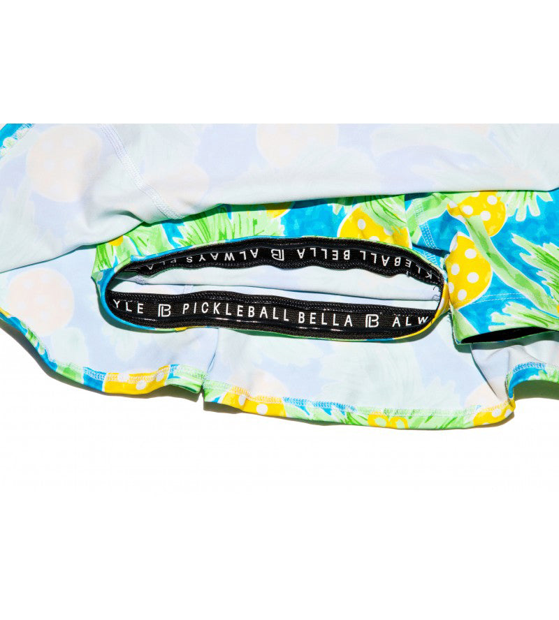 Close-up of Pickleball Bella’s PB Aline Skort Palms 1, featuring a vibrant pineapple and green leaf print, a wide waistband with white PICKLEBALL BELLA ALWAYS text, and light blue lining for comfort and style on the court.