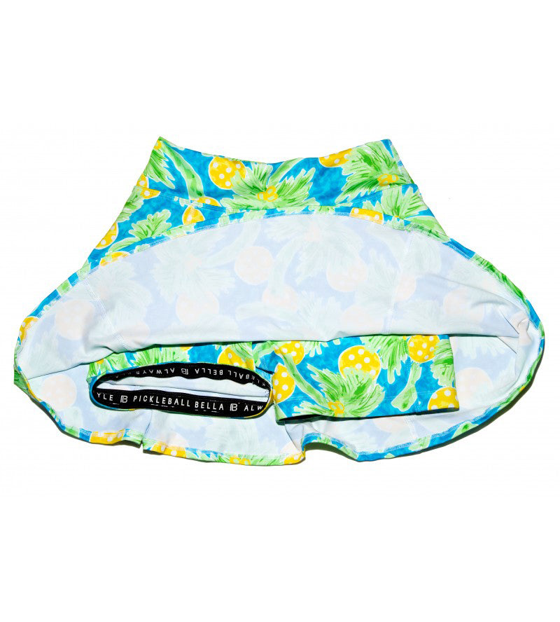 The Pickleball Bella PB Aline Skort Palms 1 is a women’s skort with a vibrant pineapple and palm print in yellow, green, and blue, featuring attached compression shorts and a wide black waistband with white “PICKLEBALL BELLA” branding.