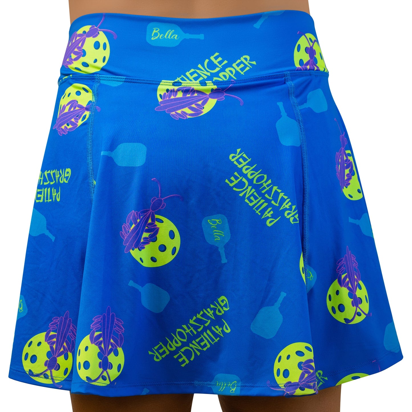 The Pickleball Bella PB Aline Skort Patience Grasshopper features a blue pickleball pattern, flared hem, built-in compression shorts, and a yellow-green “PATIENCE GRASSHOPPER” on the waistband with a small Bella logo.