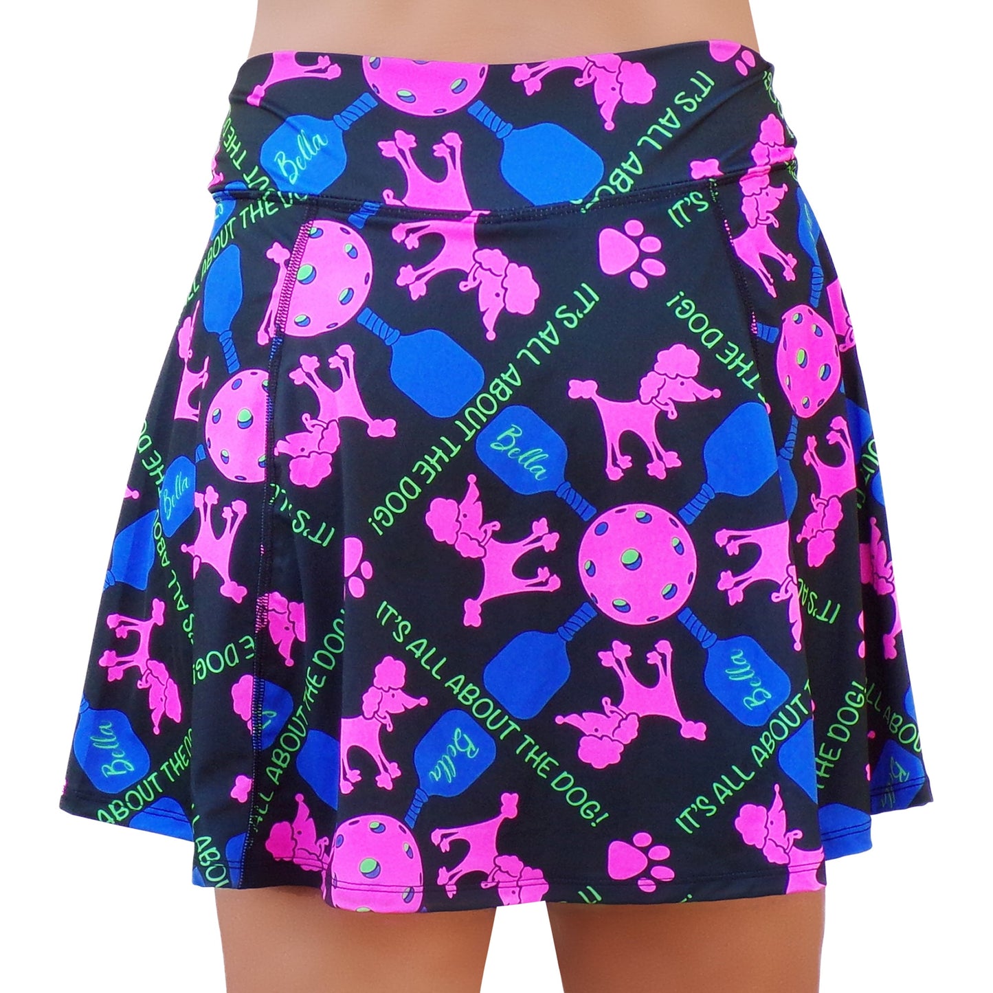 The PB Aline Skort Pickles by Pickleball Bella is a black athletic skort with a flared design, built-in compression shorts, and a vibrant print of pink poodles, tennis balls, blue “Bella” dog bones, and “ITS ALL ABOUT THE DOG!” in green.