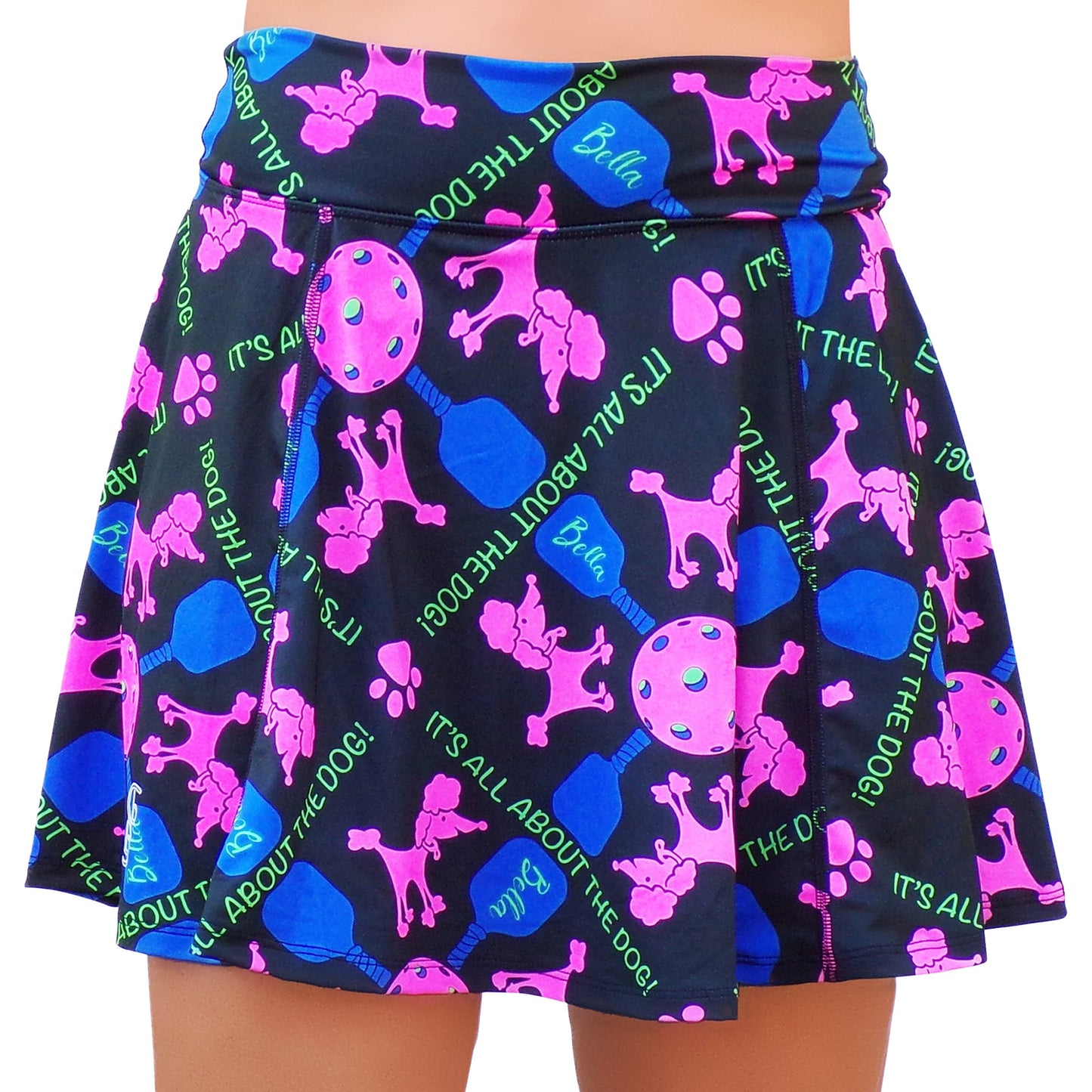 The Pickleball Bella PB Aline Skort Pickles is a flared black athletic skort with colorful pink poodles, blue dog bones, green paw prints, and lollipops. Features built-in compression shorts and "BELLA" and "ITS ALL ABOUT THE DOG!" in bright green text.