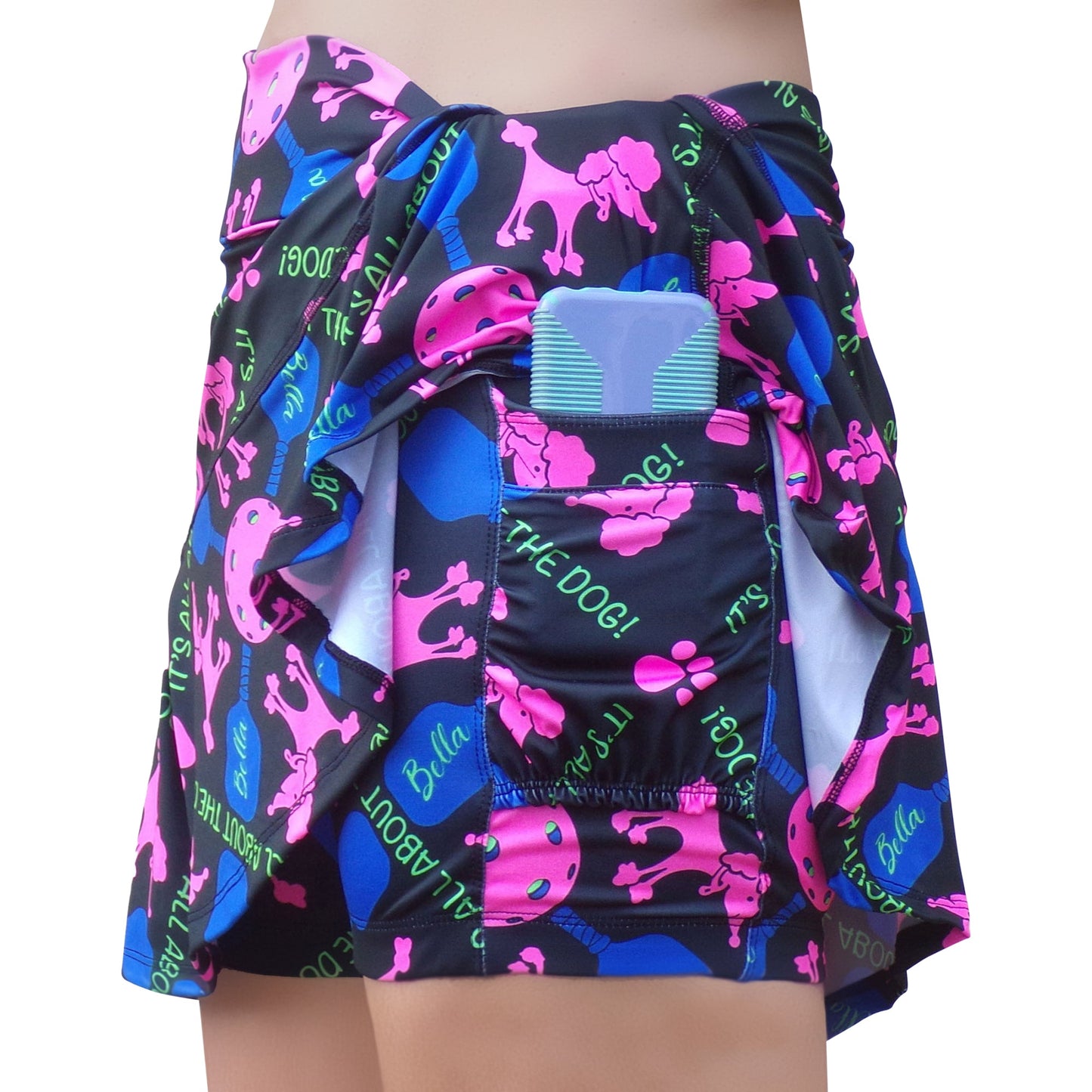 Someone wears the Pickleball Bella PB Aline Skort Pickles, featuring a playful dog-themed print with pink, blue, and green text. The skirt lifts to show built-in compression shorts with a striped pocket that holds a smartphone.