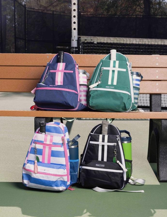 Four Fresh Pickle Teddi Pickleball Backpacks with stripe patterns—navy blue-pink, green-white, light blue-pink, and black-white—are shown by a tennis court. Two are on a brown bench, two on the ground. Each holds a water bottle.