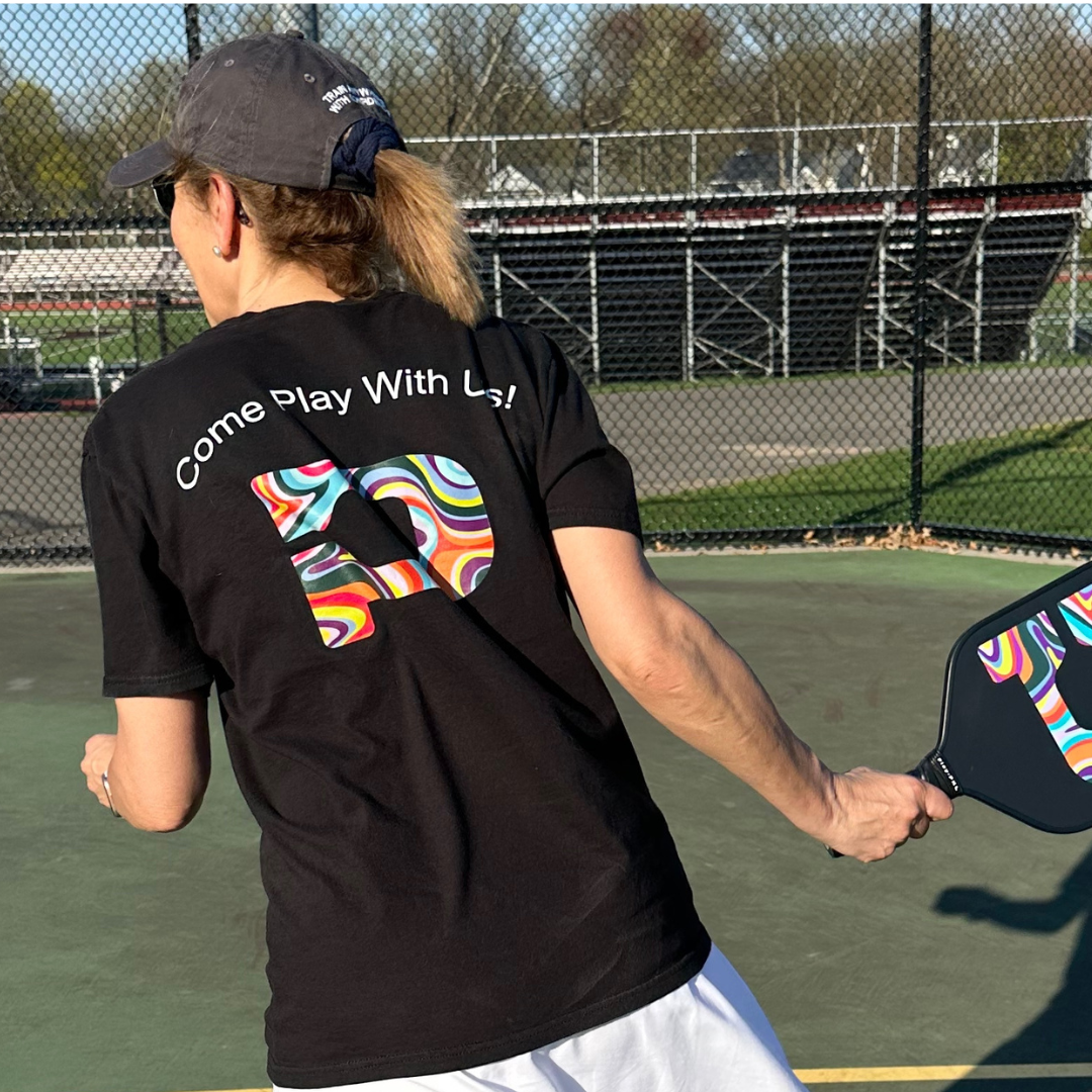 A person with a ponytail, wearing a dark cap and the Playly Store's unisex cotton "Playly - Logo T" featuring "Come Play With Us!" and a colorful patterned D on the back, plays pickleball outdoors near bleachers and fencing.