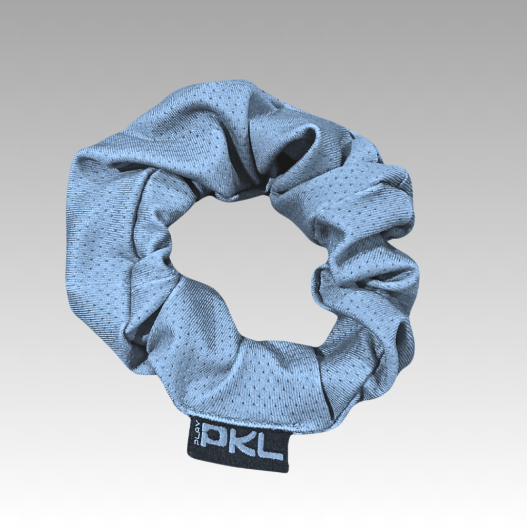 The Shanti Pickleball Hair Scrunchie by Playly Store is a light gray, perforated accessory with a black tag featuring white PKL lettering, shown against a gradient gray background.