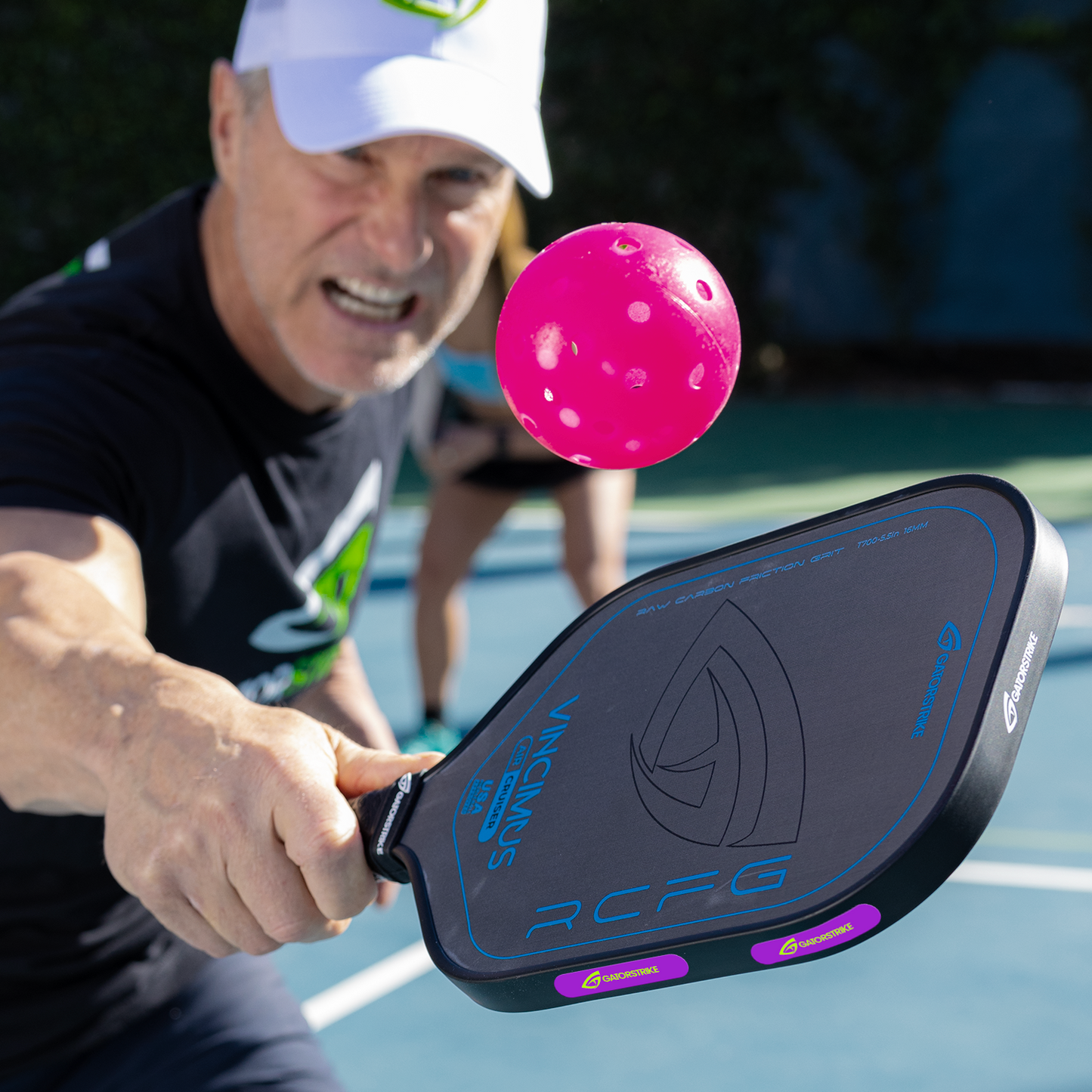 A man plays pickleball outdoors, reaching forward with a black paddle balanced using GATORSTRIKE™ Weight Tape 10 Pack by GATORSTRIKE. A bright pink ball is mid-air as the player and court blur in the background.