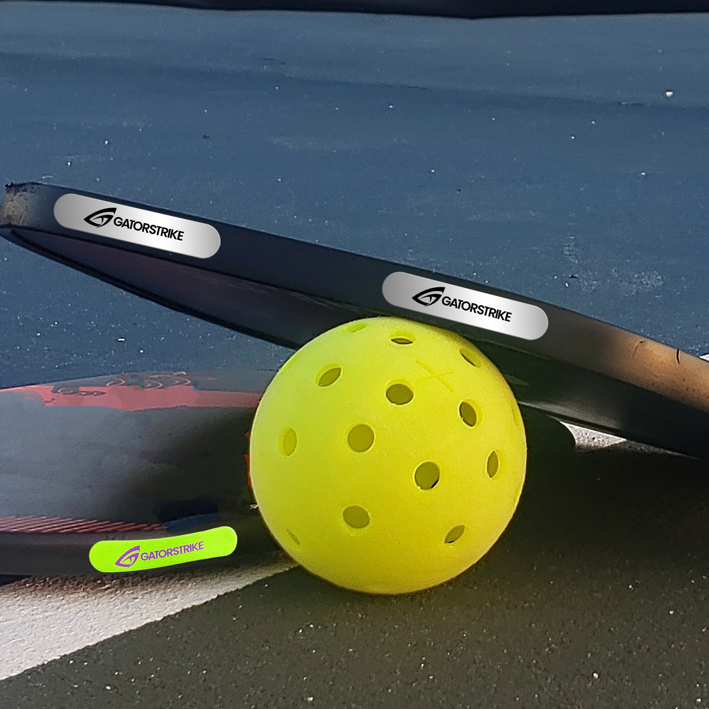 Two GATORSTRIKE™ Weight Tape rolls rest on the court—one with a white label, one gray. A bright yellow pickleball sits in front, while part of another GATORSTRIKE tape roll with a neon green label is also visible.