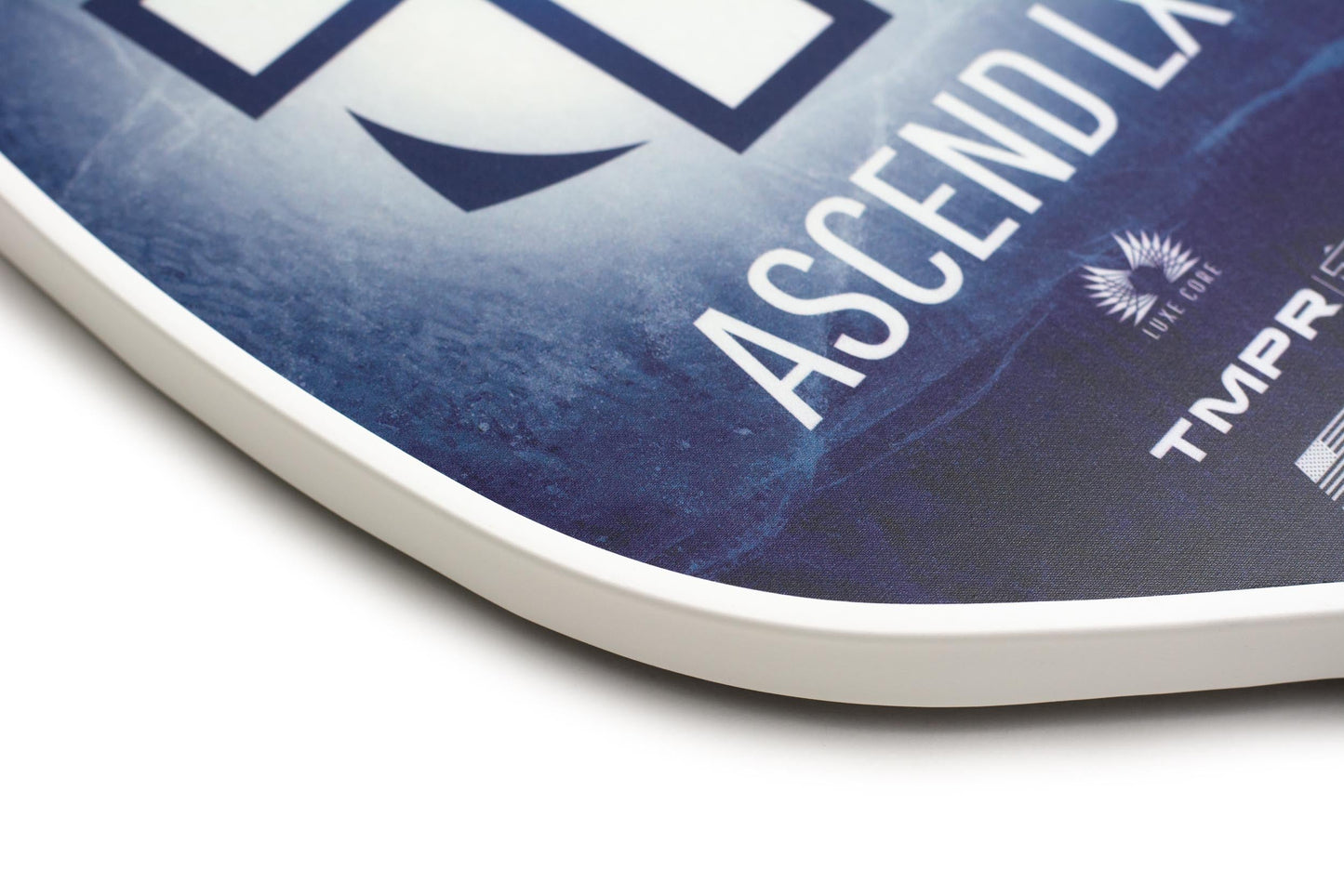 Close-up of the curved corner of the TMPR Sports Ascend LX | Indigo Ice pickleball paddle with a blue and white design, feathered emblem, and logos. The long handle is visible as the background fades into deep blue.