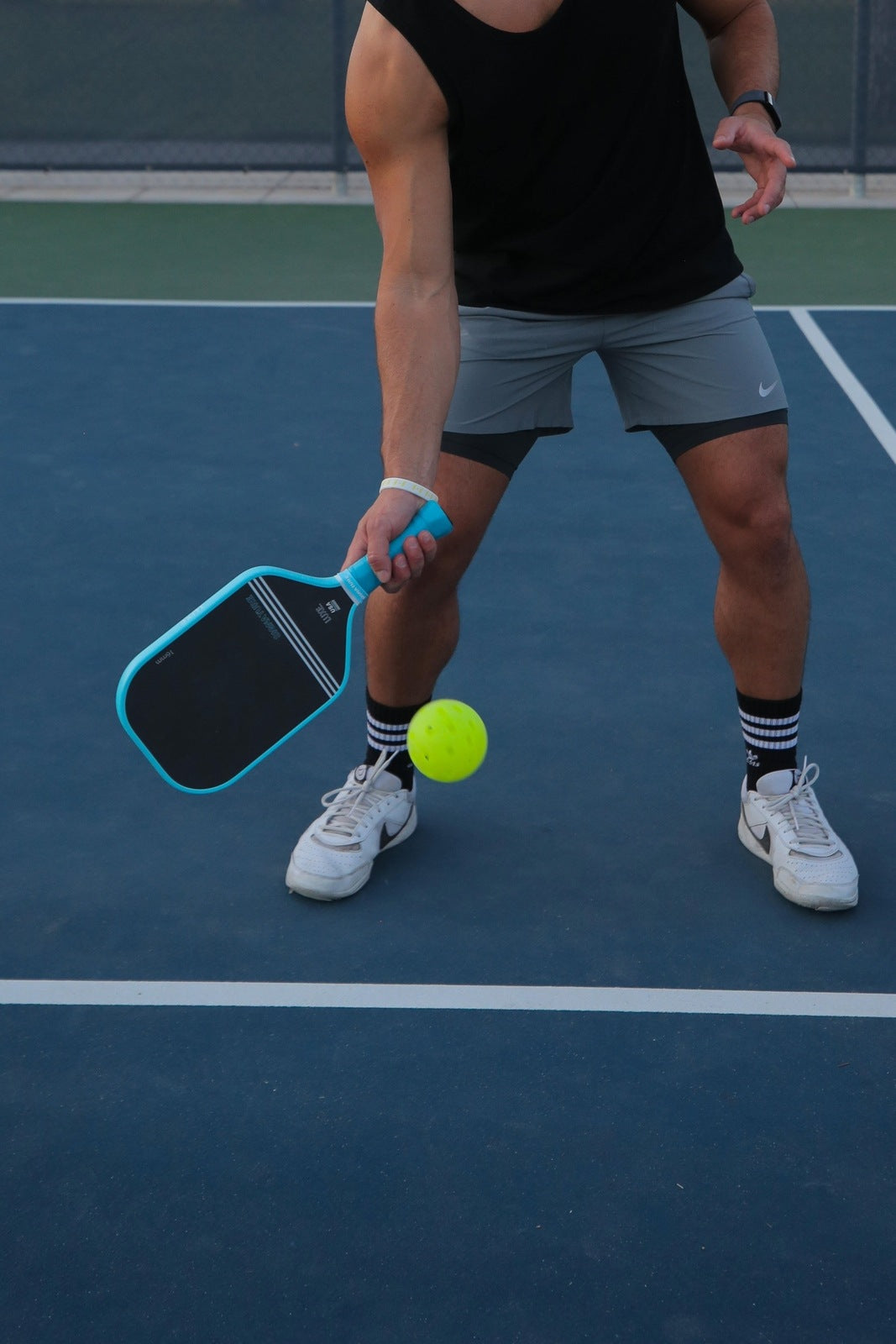 Wearing striped black socks and white shoes, a player uses the LUXE Pickleball Signature paddle to hit a yellow ball on an outdoor dark blue court with white lines. Only their torso and legs are visible in athletic wear.