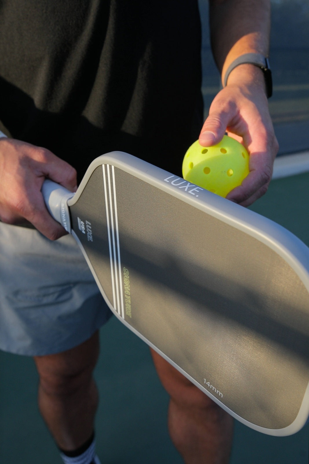 A person in a black shirt and light shorts holds a gray LUXE Pickleball Signature paddle in one hand and a yellow perforated pickleball in the other, ready to serve on an outdoor court lit by natural sunlight.