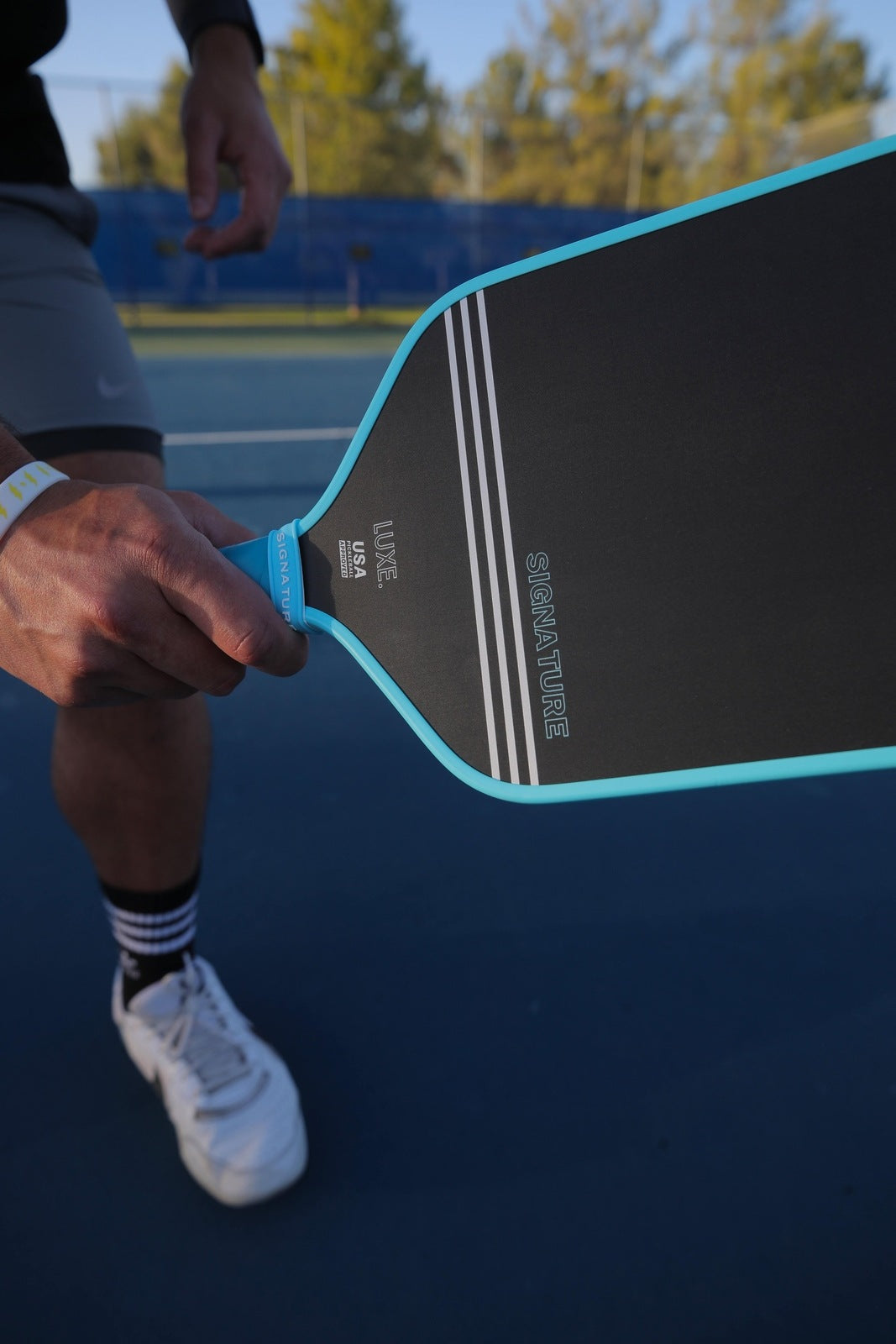 On an outdoor court, a person holds the LUXE Pickleball Signature paddle—black and blue carbon fiber—while wearing white shoes, striped socks, gray shorts, and a pale wristband. Trees and a blue net are visible in the background.
