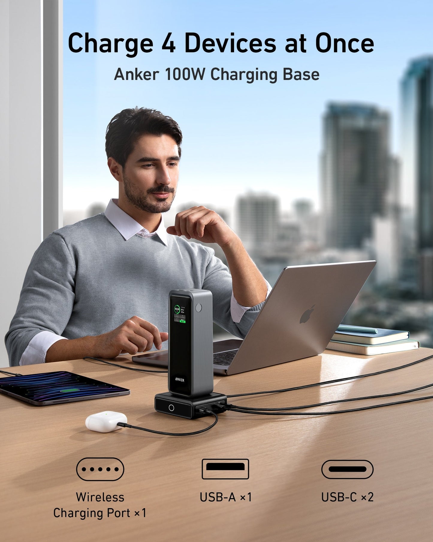 A man works at a desk with a laptop, tablet, and mouse, using the Anker Prime 27,650mAh Power Bank (250W) and 100W Charging Base to charge four devices at once via wireless pad, USB-A, and two USB-C ports. City buildings are seen through large windows behind him.