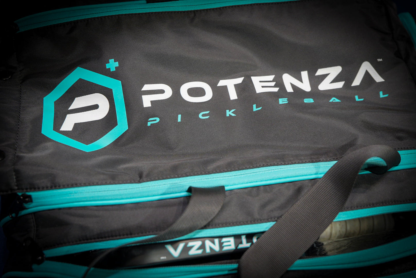 The Potenza Pickleball Tournament Paddle Bag is black with teal zippers and accents, featuring a bold white and teal logo with a stylized P in a teal hexagon on the front pocket. A paddle peeks out, highlighting its protective design.