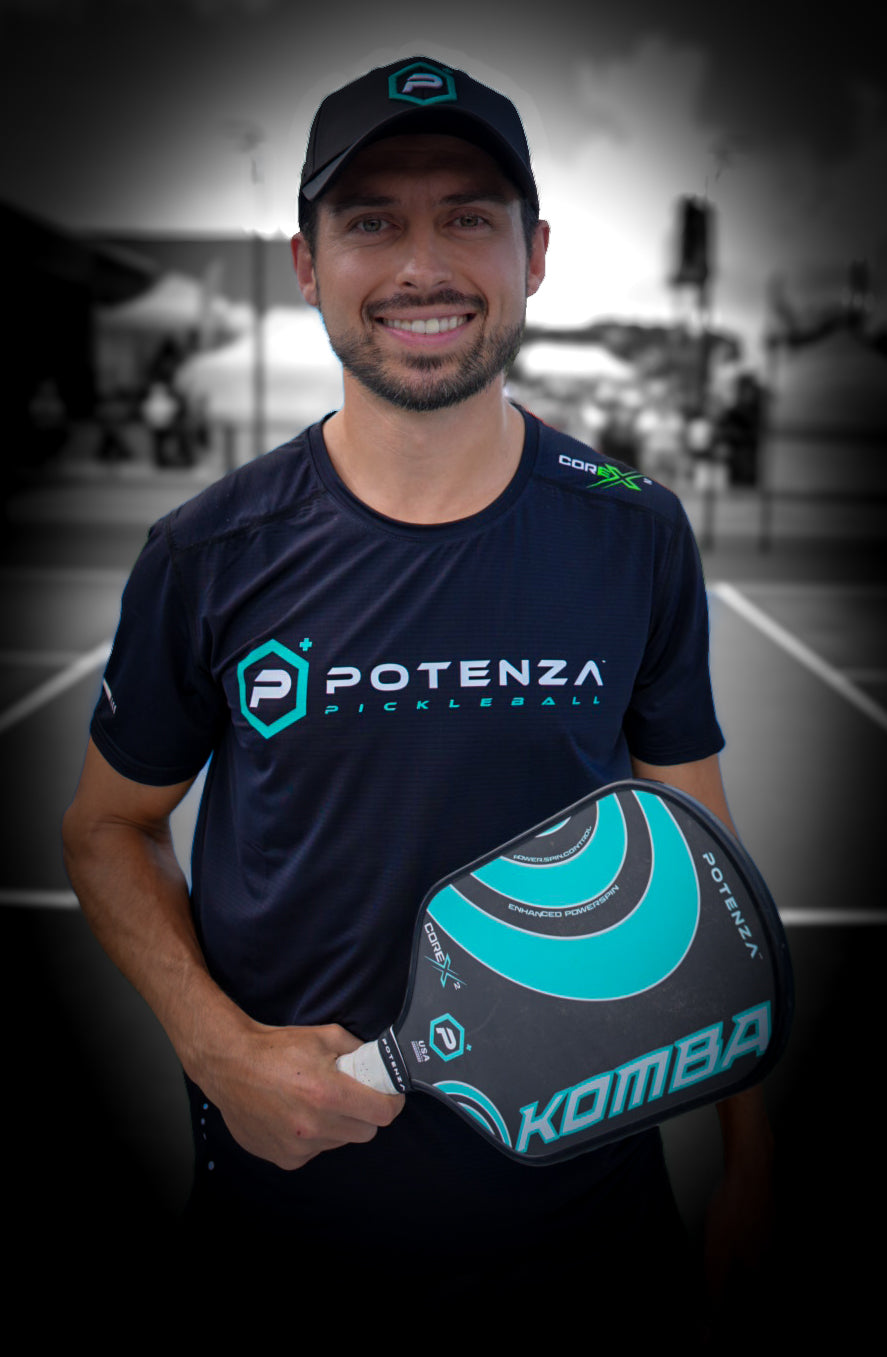 A man in a Potenza Pickleball Quick-Dry Performance T-Shirt and cap smiles at the camera, holding a turquoise and black Komba pickleball paddle on an outdoor court with blurred nets and players behind him.