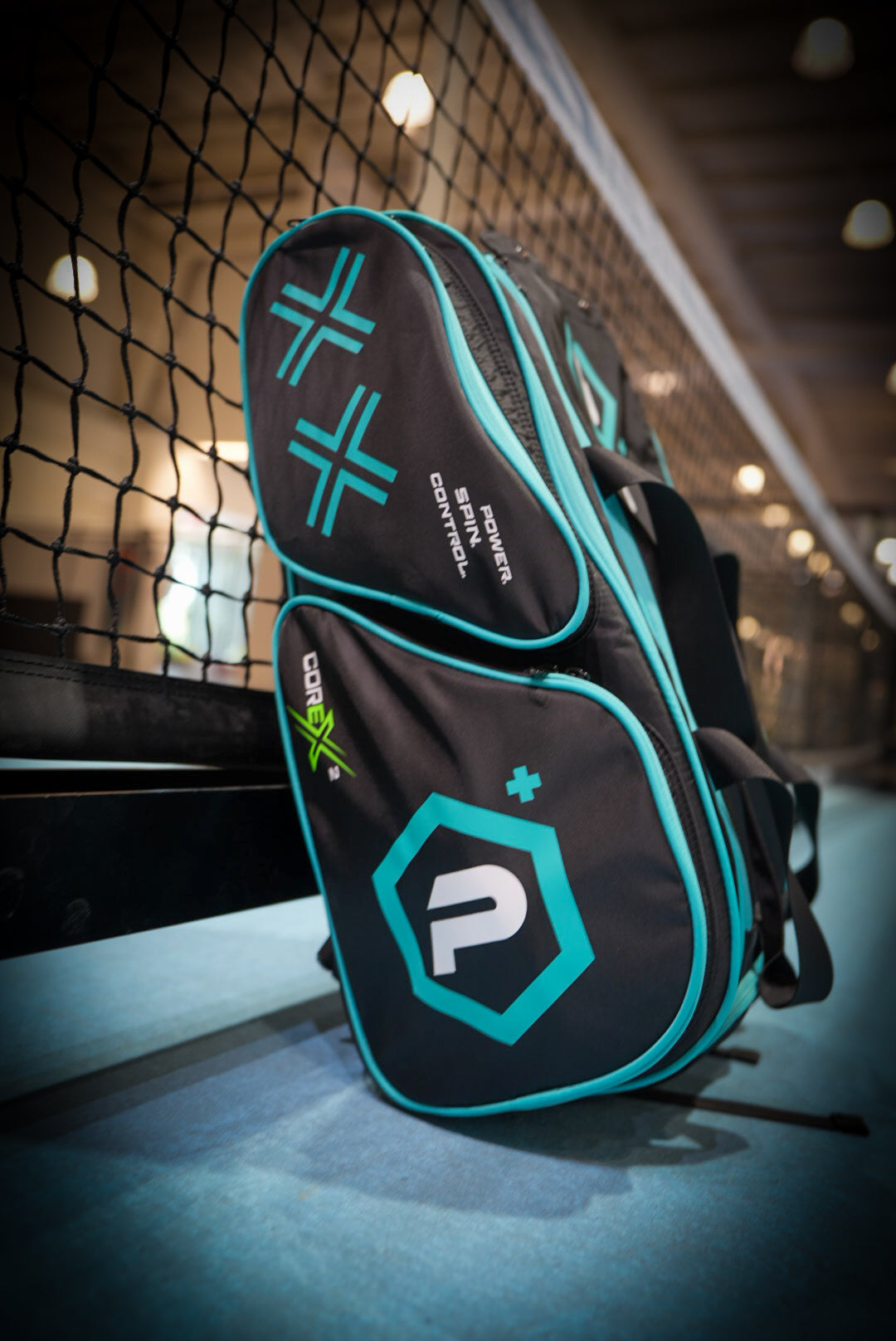 The Potenza Pickleball Tournament Paddle Bag, in black and teal with geometric logos, leans against an indoor court net. Featuring paddle protection, multiple compartments, and bold "Control" and "Power" text, it stands out on the blue surface.
